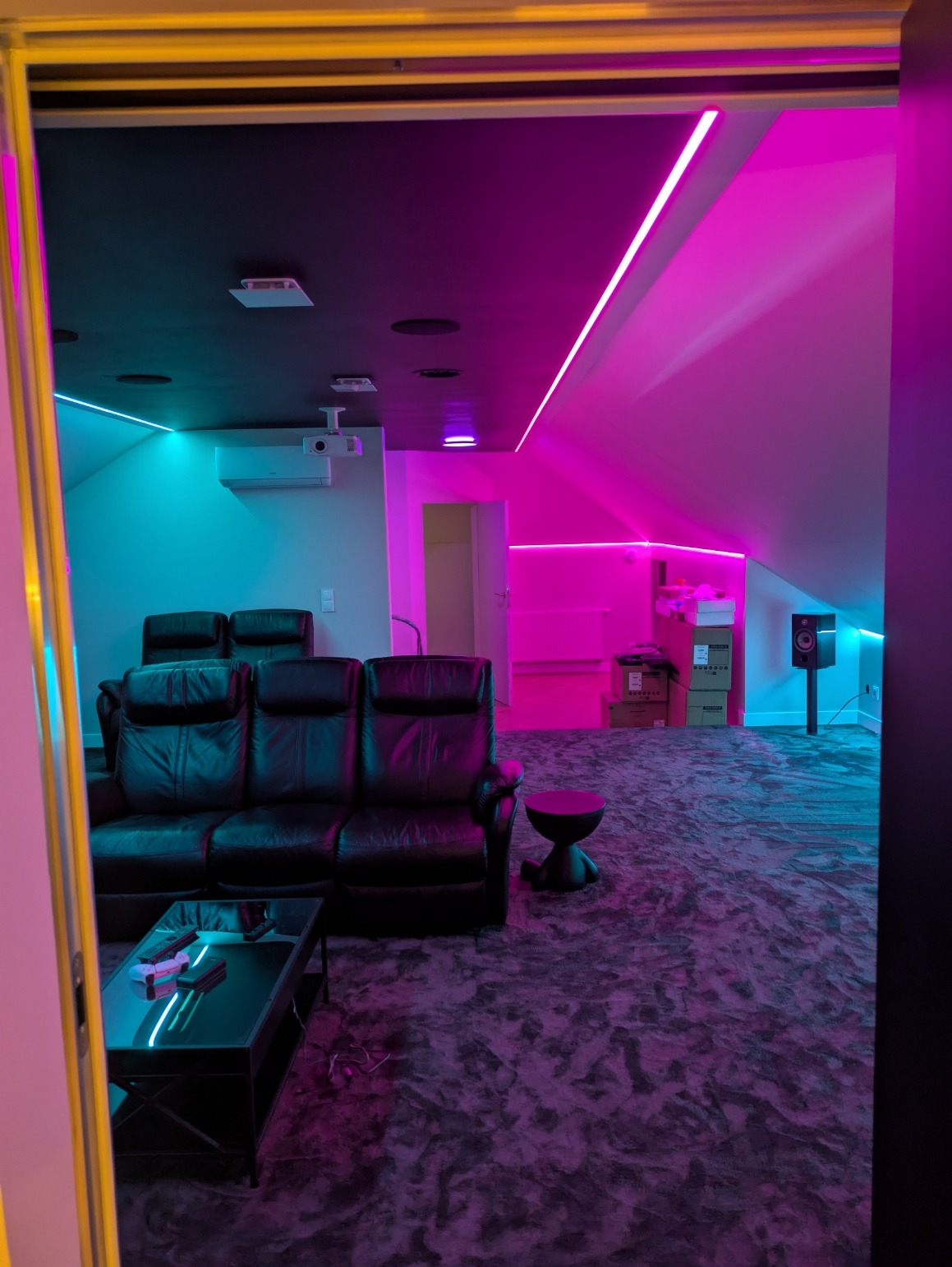 Kino domowe w pokoju na poddaszu z czarną skórzaną sofą, stolikiem i oświetleniem LED w kolorach różu i turkusu. Widoczny projektor i głośnik.
