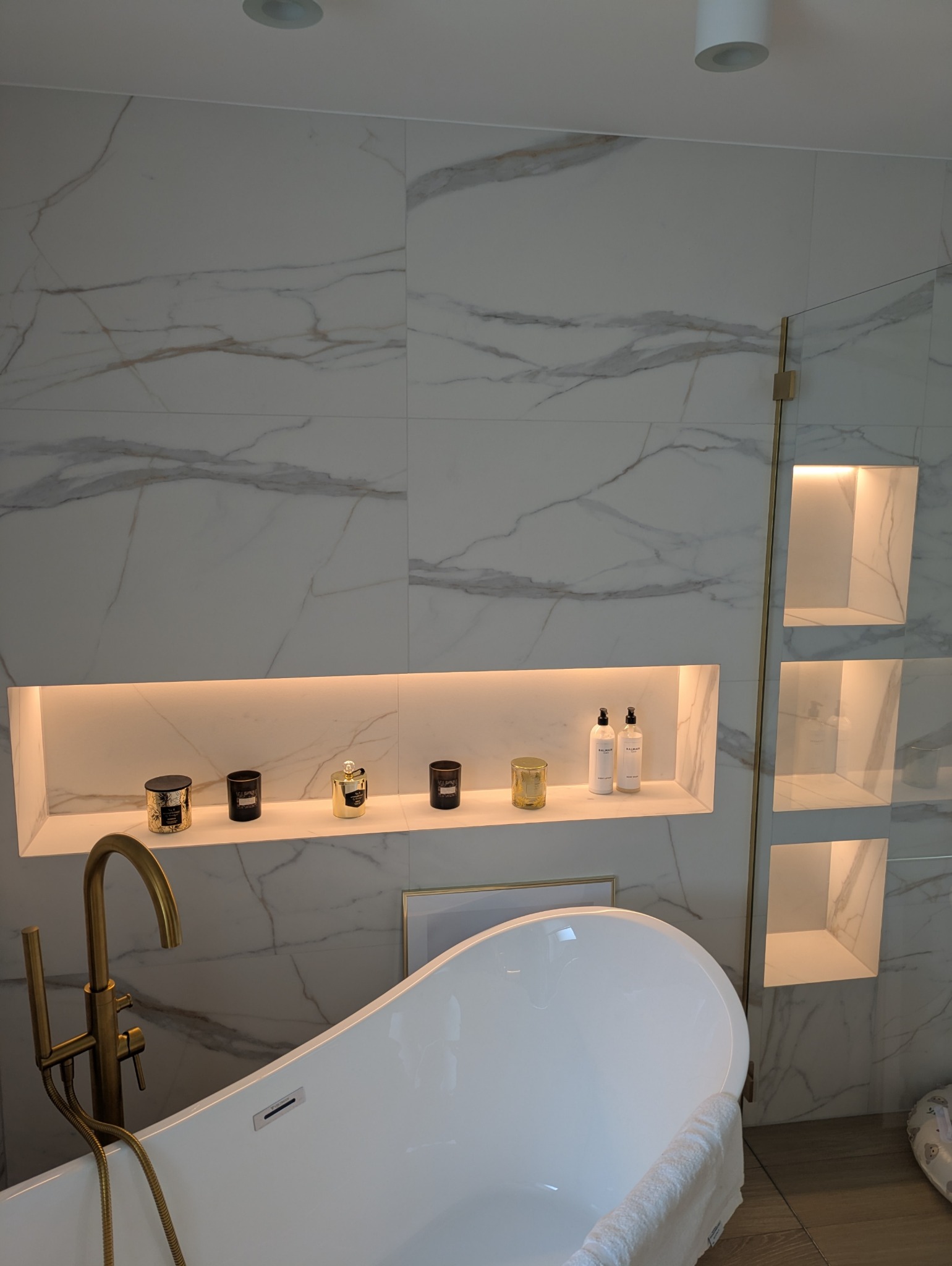 Nowoczesna łazienka z wanną wolnostojącą, złotą armaturą i podświetlanymi wnękami w marmurowej ścianie. Minimalistyczny design z akcentami luksusu.
