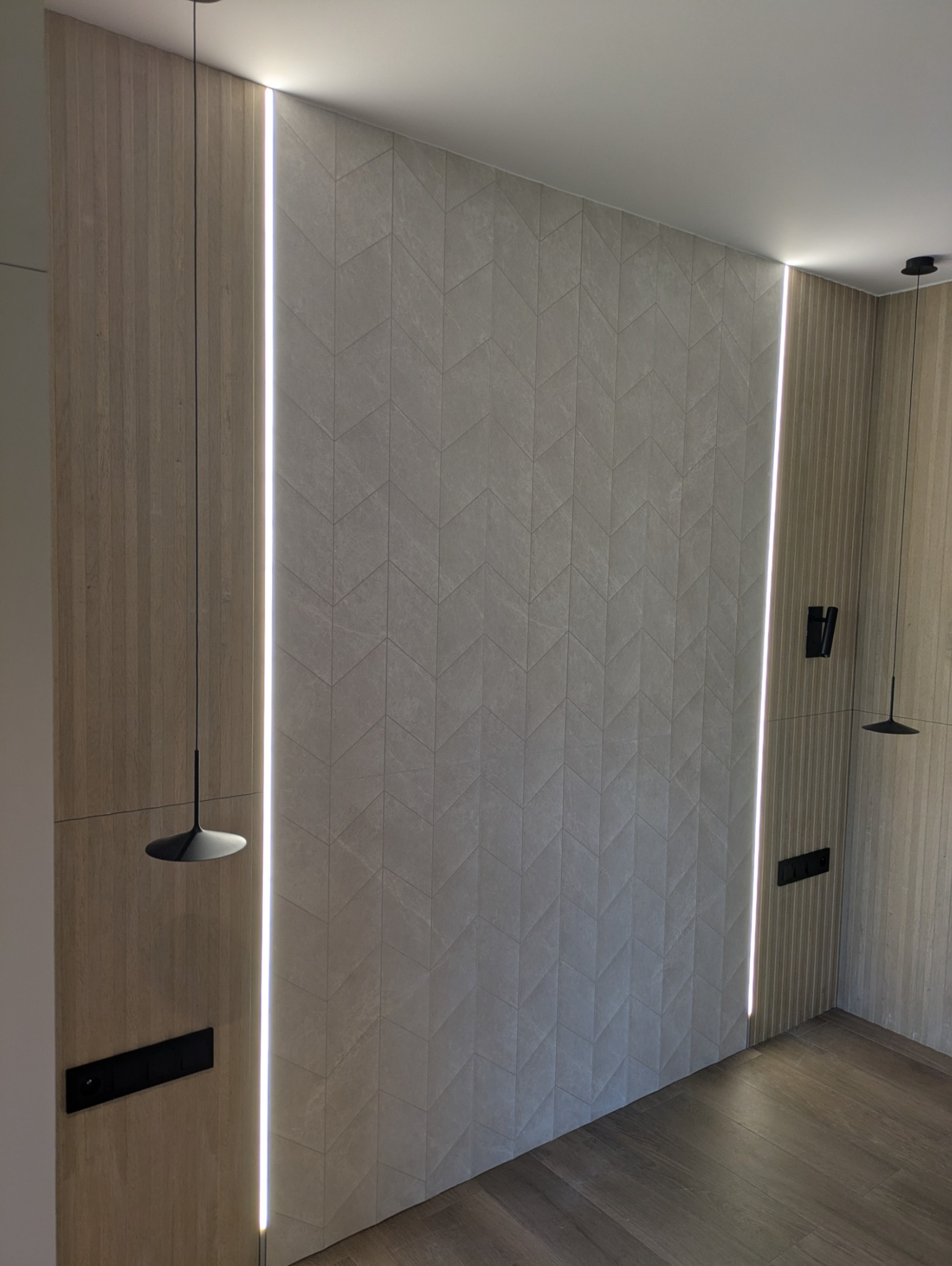 Ściana z dekoracyjną, podświetlaną listwą LED, wykończona panelami drewnopodobnymi i płytkami w jodełkę. Widoczne minimalistyczne lampy wiszące i gniazdka.