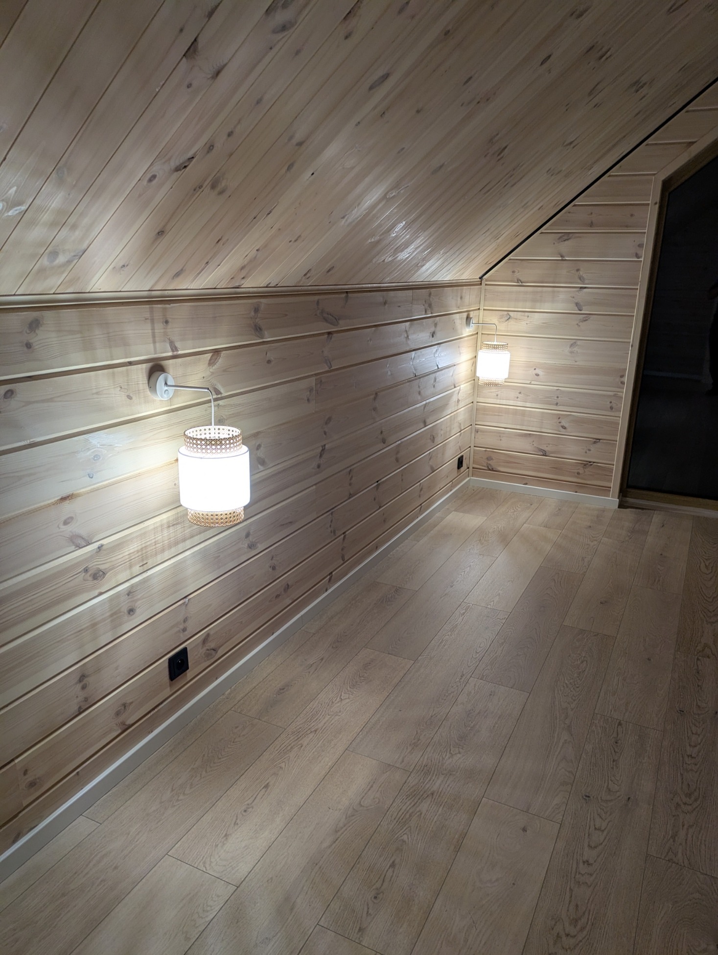 Drewniane wnętrze poddasza z panelami ściennymi i podłogą w odcieniach beżu. Dwa kinkiety z wiklinowym wzorem oświetlają przestrzeń. Widoczne gniazdko elektryczne.