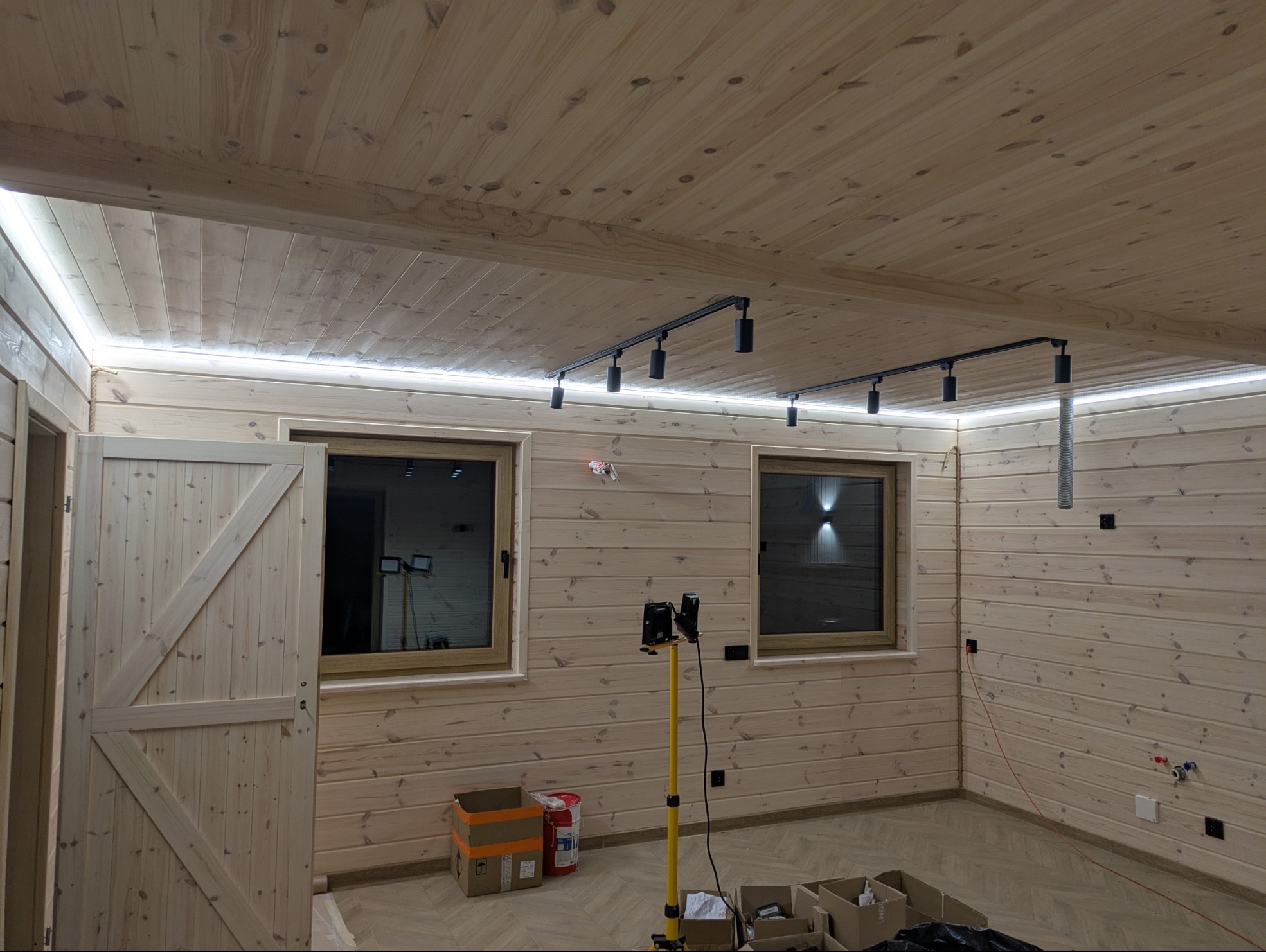 Drewniane wnętrze z oświetleniem LED podsufitowym i reflektorami na szynoprzewodzie. Widoczne okna, drzwi stodołowe i narzędzia na podłodze.