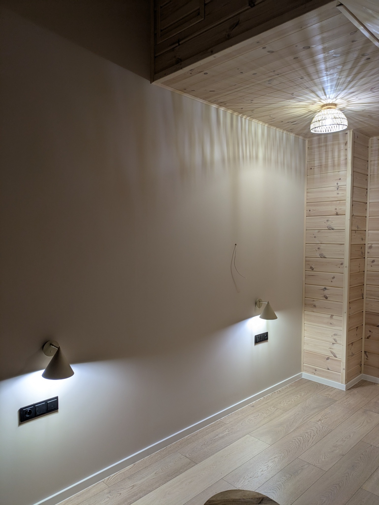 Jasne wnętrze z drewnianym sufitem i ścianami, oświetlone kinkietami i lampą sufitową. Widoczne gniazdka elektryczne i wystający kabel.