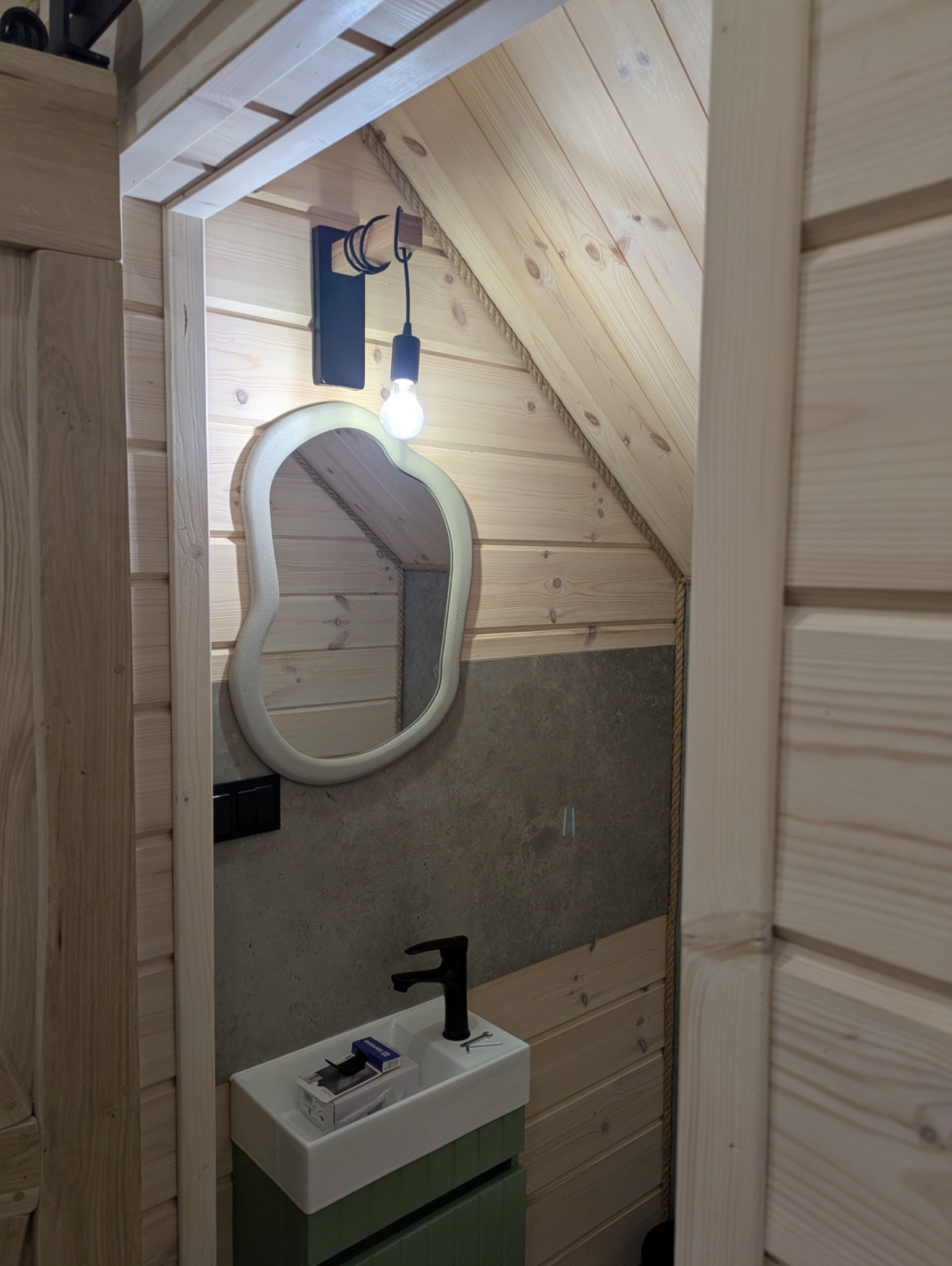 Drewniane wnętrze łazienki na poddaszu z asymetrycznym lustrem, czarną lampą i umywalką. Ściany wykończone boazerią i szarą płytą. Widoczne detale wykończeniowe.