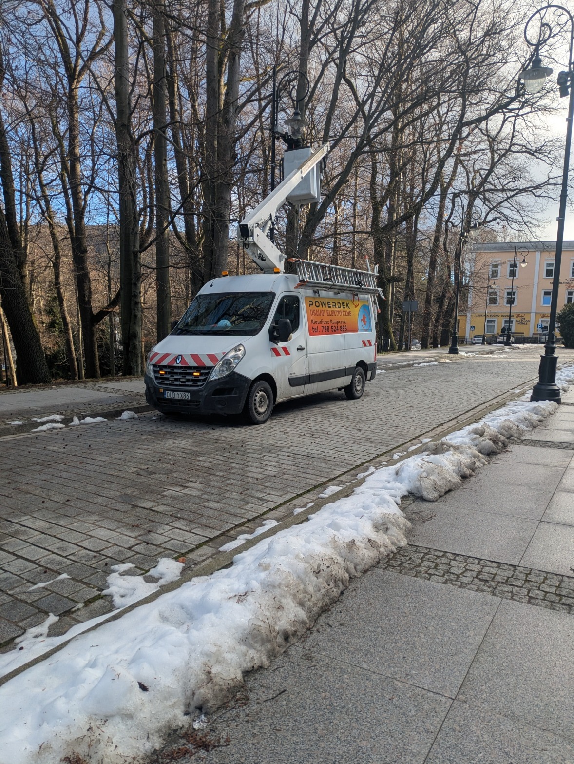Pracownik w podnośniku koszowym naprawia oświetlenie uliczne. Biały samochód z logo firmy elektrycznej zaparkowany na brukowanej ulicy w zimowy dzień.