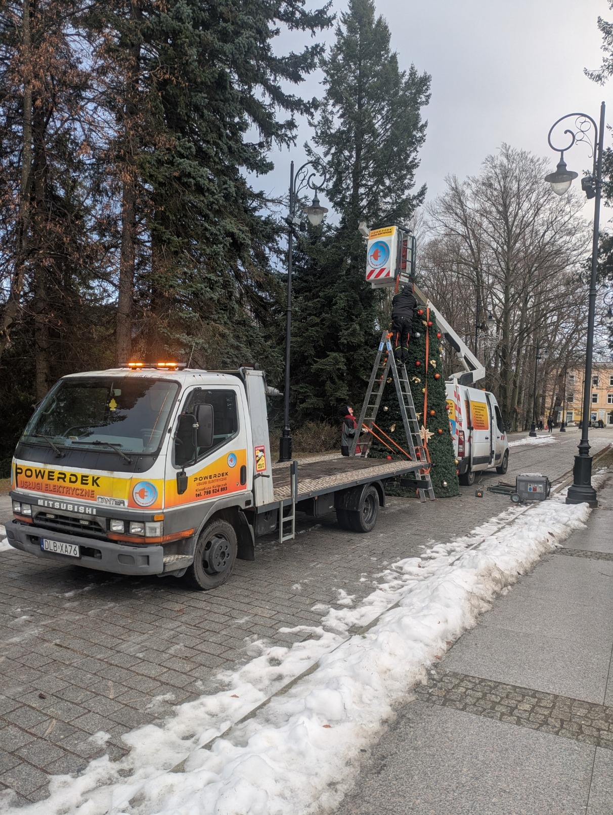 Montaż dekoracji świątecznych na zewnątrz przez firmę POWERDEK. Pracownicy na podnośniku i drabinie dekorują choinkę. Na pierwszym planie samochód dostawczy firmy.