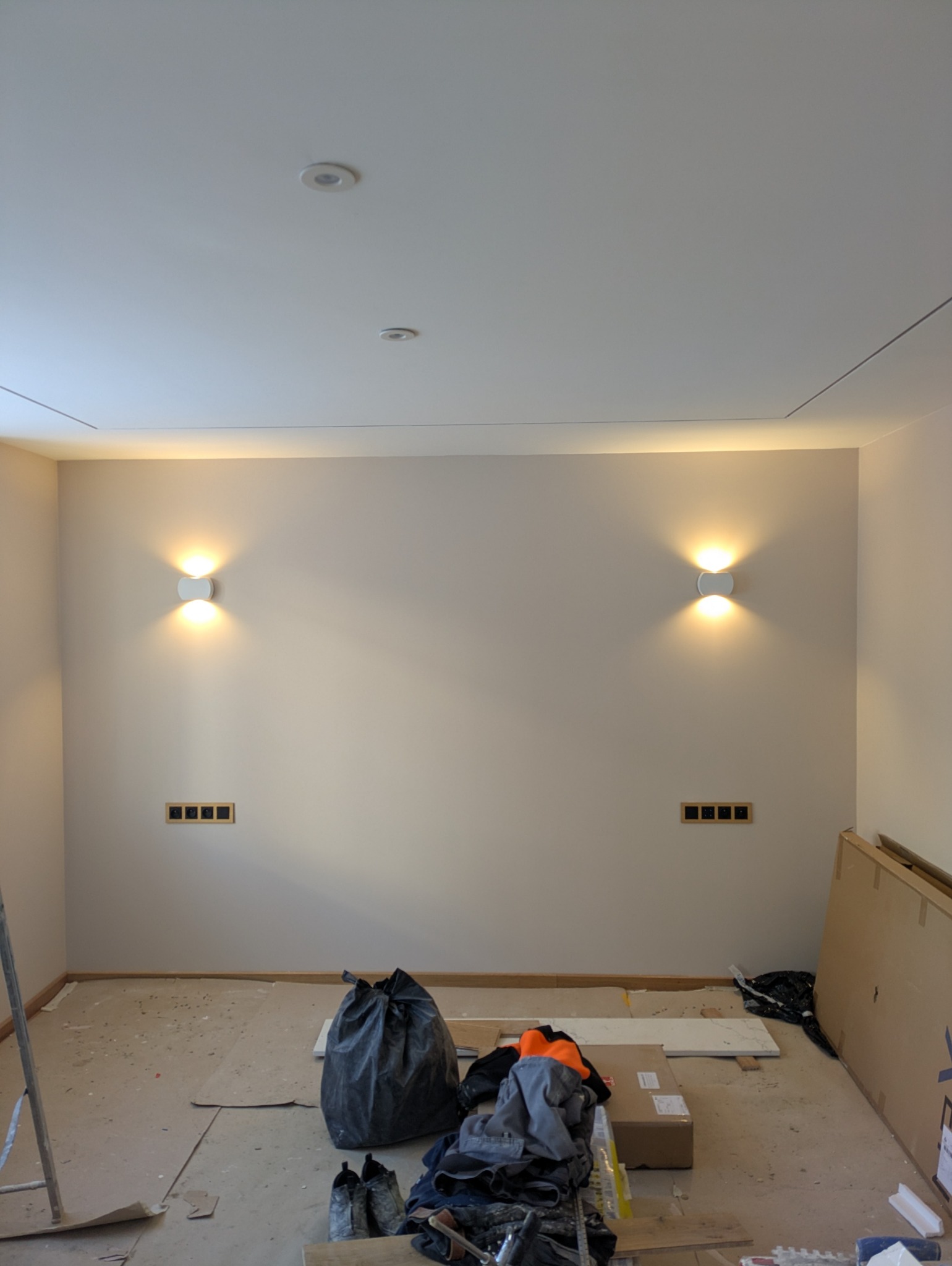 Pokój w trakcie remontu: zamontowane kinkiety ścienne, puszki elektryczne, podłoga zabezpieczona kartonem, widoczne narzędzia i worki z odpadami budowlanymi.