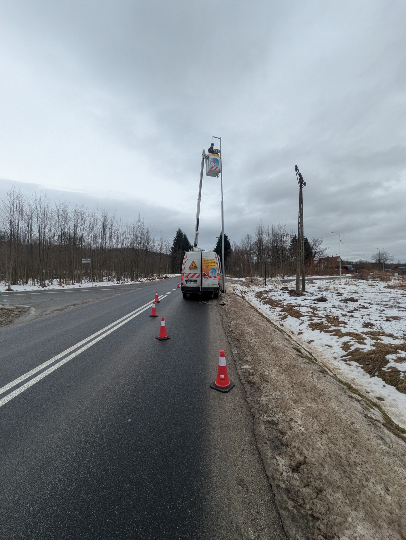 Pracownik w podnośniku koszowym naprawia oświetlenie uliczne. Na drodze ustawione pachołki ostrzegawcze, w tle las i pochmurne niebo. Widoczny samochód serwisowy.