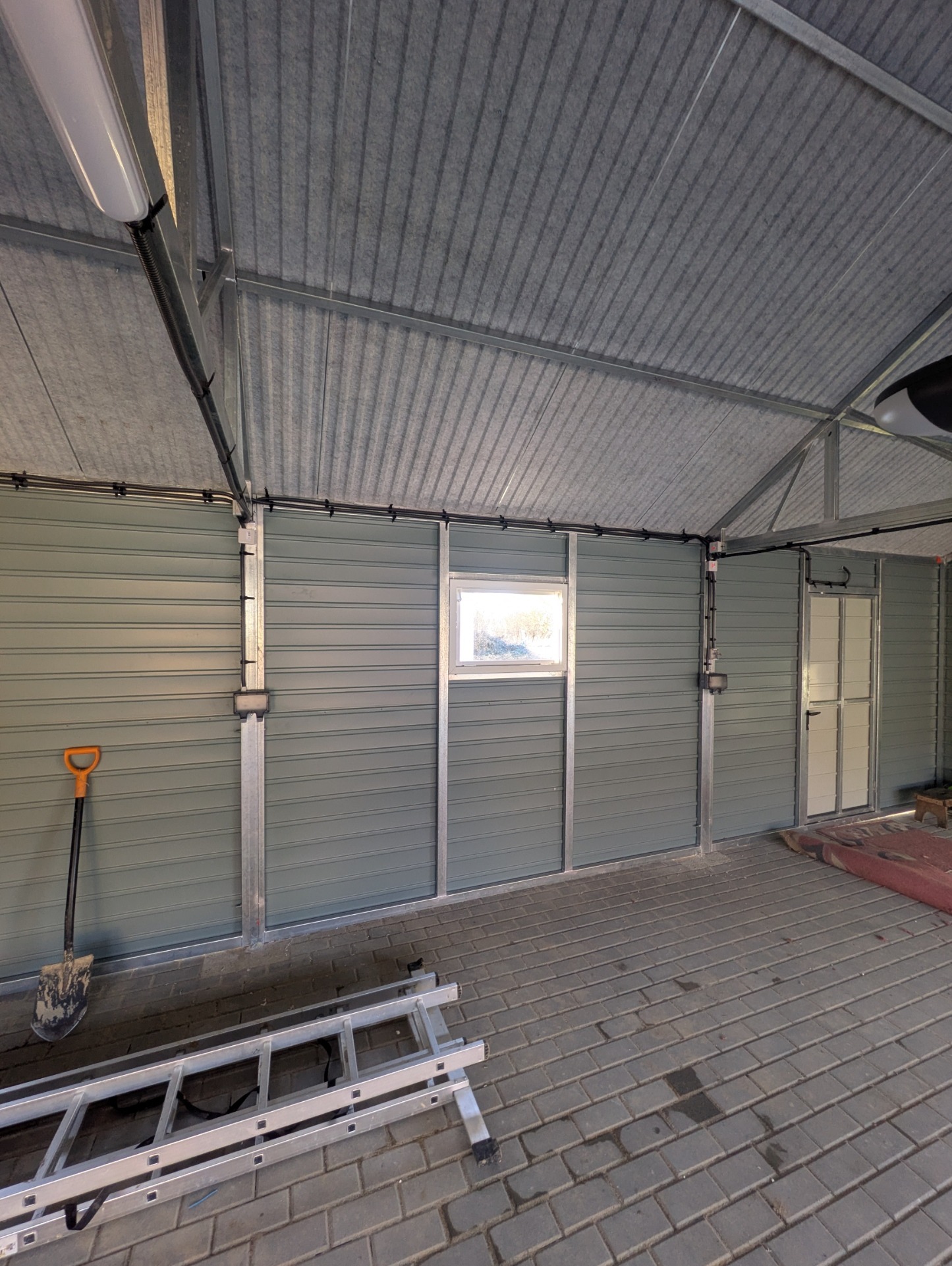 Wnętrze garażu z szarą blachą trapezową na ścianach i suficie, widoczne instalacje elektryczne, drabina i łopata. Oświetlenie jarzeniowe.