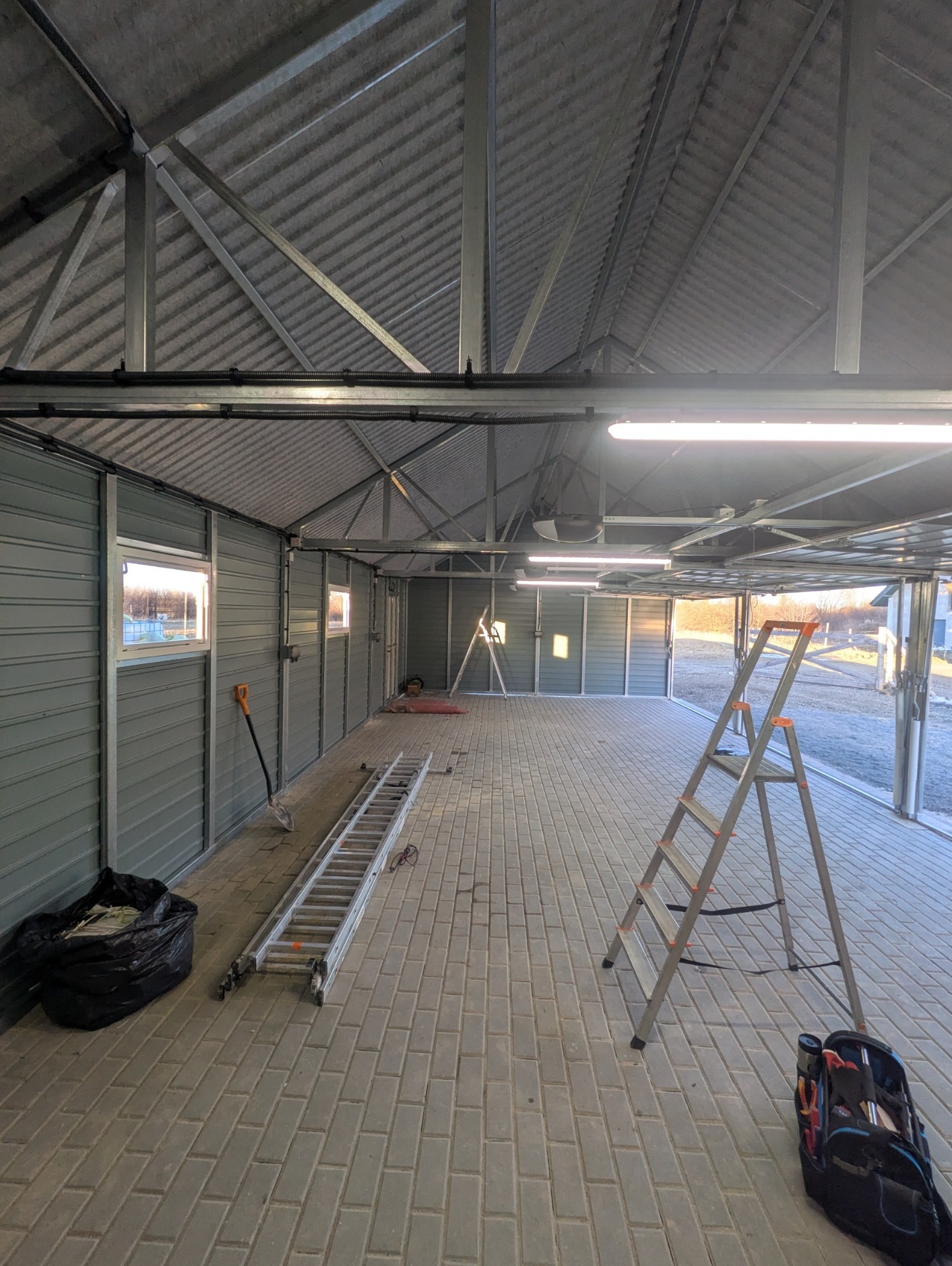 Wnętrze garażu z metalową konstrukcją dachu i ścian, widoczne drabiny, narzędzia i oświetlenie. Świecące lampy sufitowe.