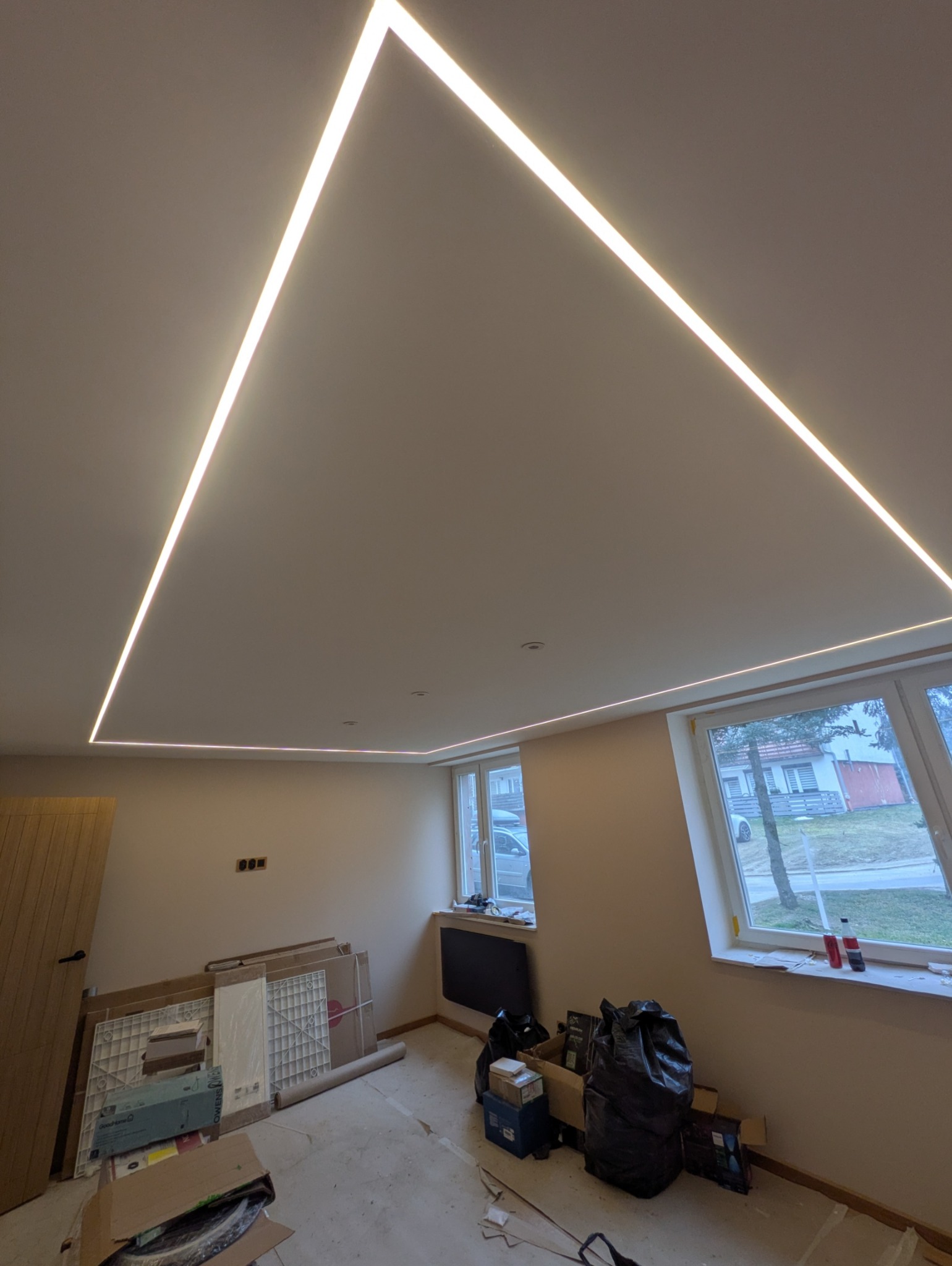 Nowoczesne oświetlenie sufitowe LED w kształcie kwadratu, w jasnym pomieszczeniu z dwoma oknami. Widoczne materiały budowlane i kartony na podłodze.