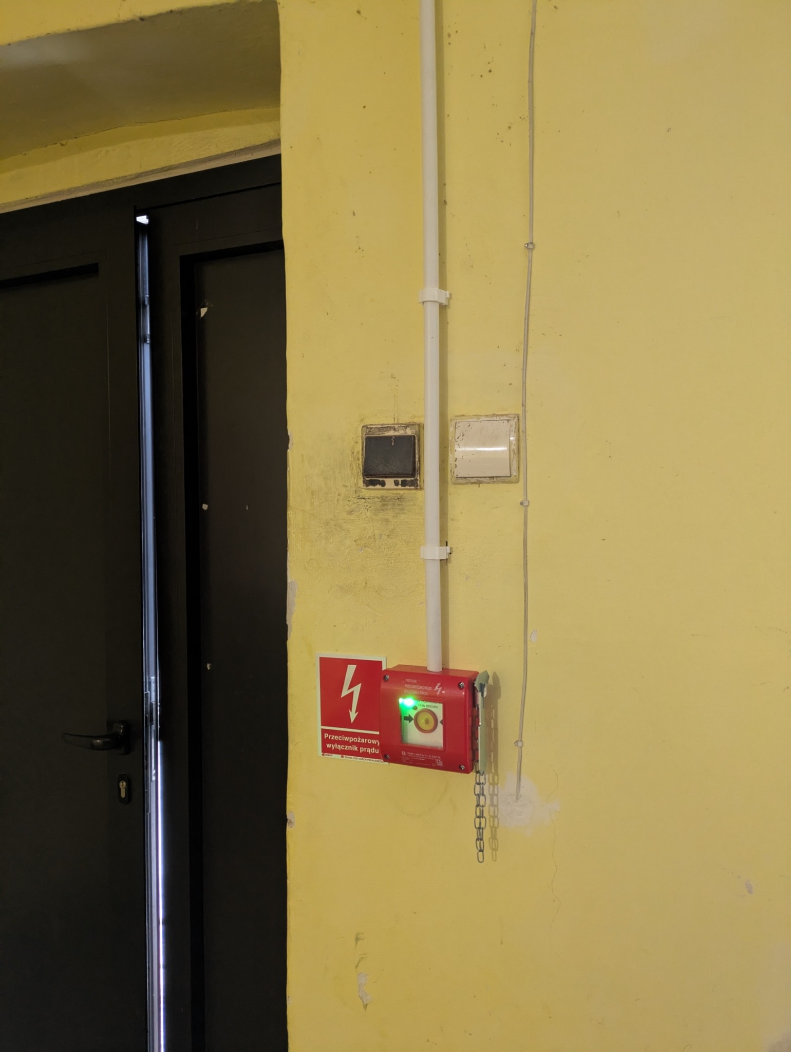Czerwony, przeciwpożarowy wyłącznik prądu z zieloną diodą, zamontowany na żółtej ścianie obok czarnych drzwi. Widoczne przewody i elementy instalacji.