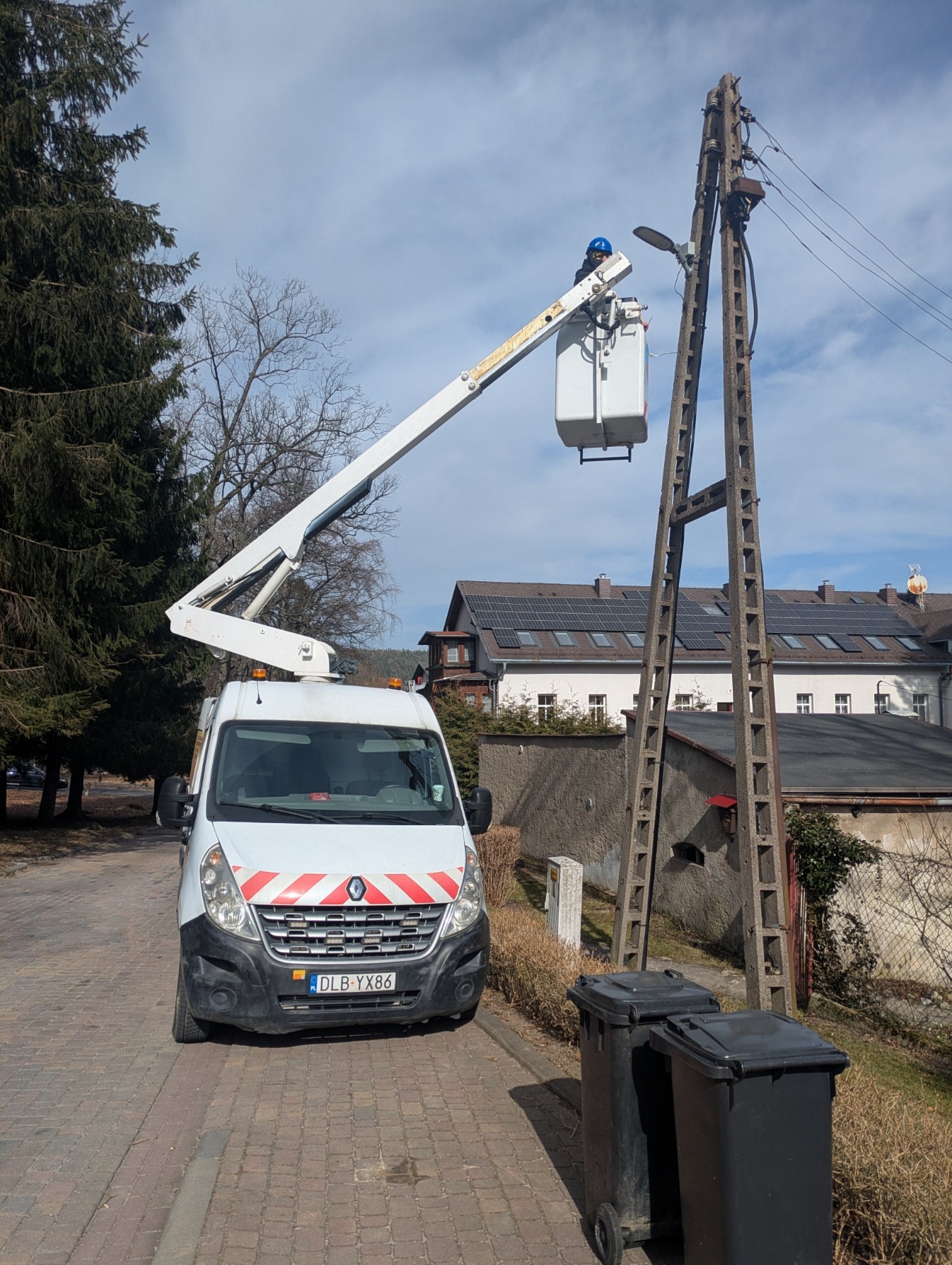 Pracownik w podnośniku koszowym przy betonowym słupie energetycznym. Biały samochód z podnośnikiem na tle zabudowań i drzew. Dwie czarne beczki na śmieci na pierwszym planie.