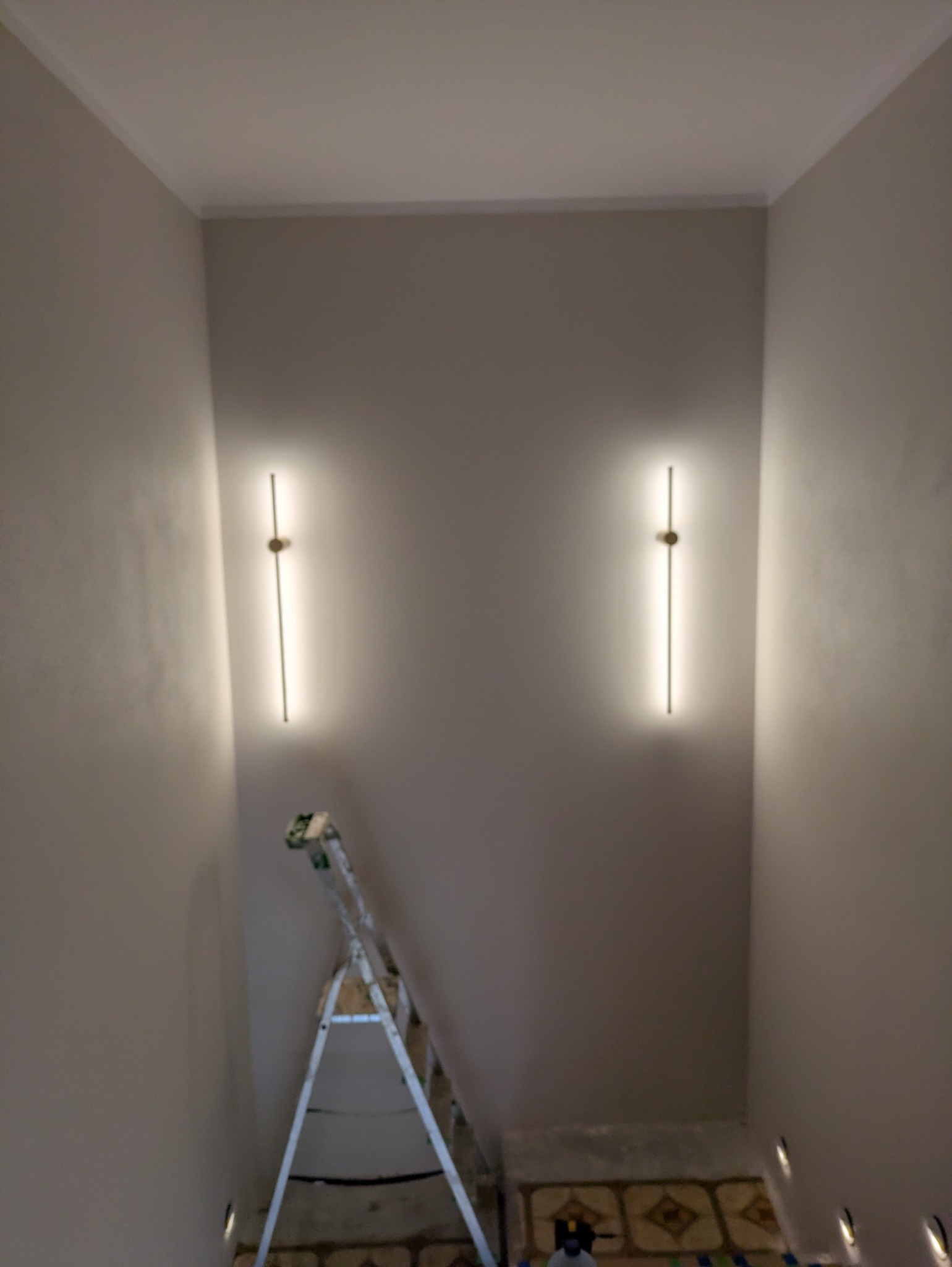 Dwa nowoczesne, pionowe kinkiety LED zamontowane na ścianie w korytarzu. Pod nimi rozstawiona drabina aluminiowa. Widoczne oświetlenie podłogowe.
