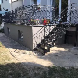 Wykonana z kostki brukowej ścieżka prowadzi do schod&oacute;w z metalową balustradą, balkon z ozdobną balustradą i donicami z kwiatami.