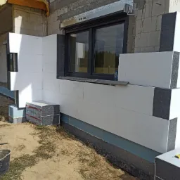 Ocieplanie elewacji domu jednorodzinnego styropianem grafitowym, widoczne okno z roletą zewnętrzną Krispol, przygotowane pakiety styropianu na ziemi.