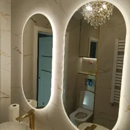 Nowoczesna łazienka z dwoma podświetlanymi lustrami o owalnym kształcie, złotą armaturą i marmurowymi płytkami na ścianach, w odbiciu widoczny żyrandol kryształowy i toaleta.