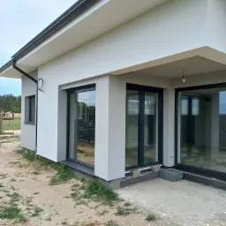 Nowoczesny dom jednorodzinny z jasną elewacją, dużymi przesuwnymi oknami z czarnymi ramami i widocznymi elementami instalacji na ścianie zewnętrznej, teren wokół domu w trakcie zagospodarowywania.