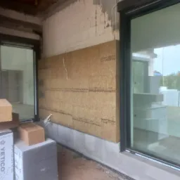 Fragment ściany zewnętrznej budynku w trakcie ocieplania wełną mineralną, widoczne okno z czarną ramą i ułożone obok bloki izolacyjne YETICO.