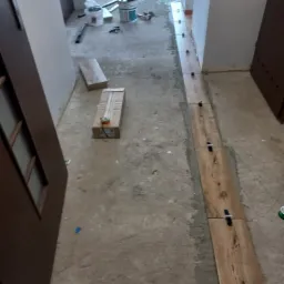 Korytarz w trakcie układania podłogi z płytek imitujących drewno, widoczne krzyżyki dystansowe, poziomica, wiadro z klejem i narzędzia glazurnicze.
