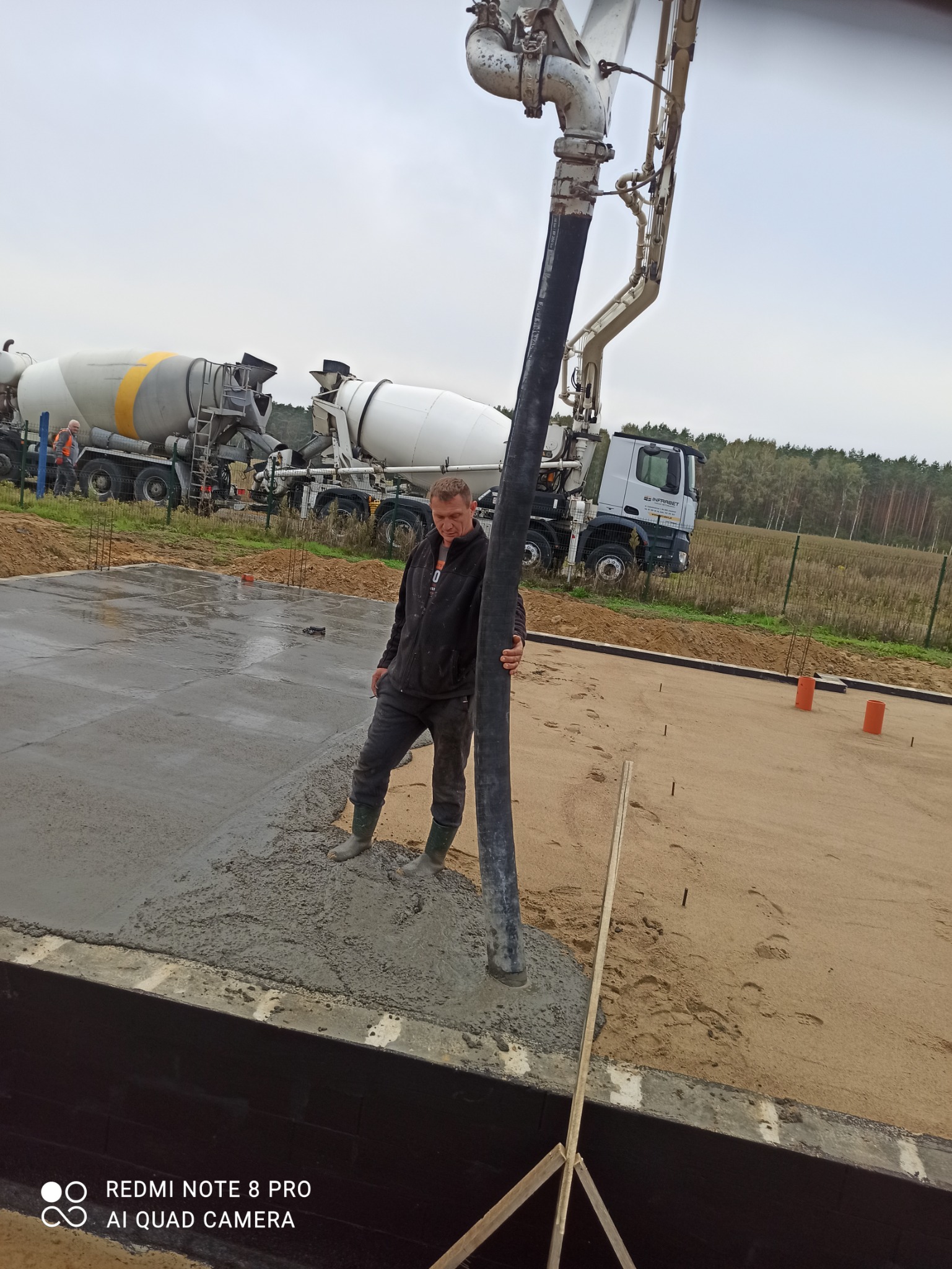 Pracownik budowlany w gumowych butach kieruje wylewką betonu z pompy na przygotowany fundament; w tle dwie betoniarki i ogrodzenie.