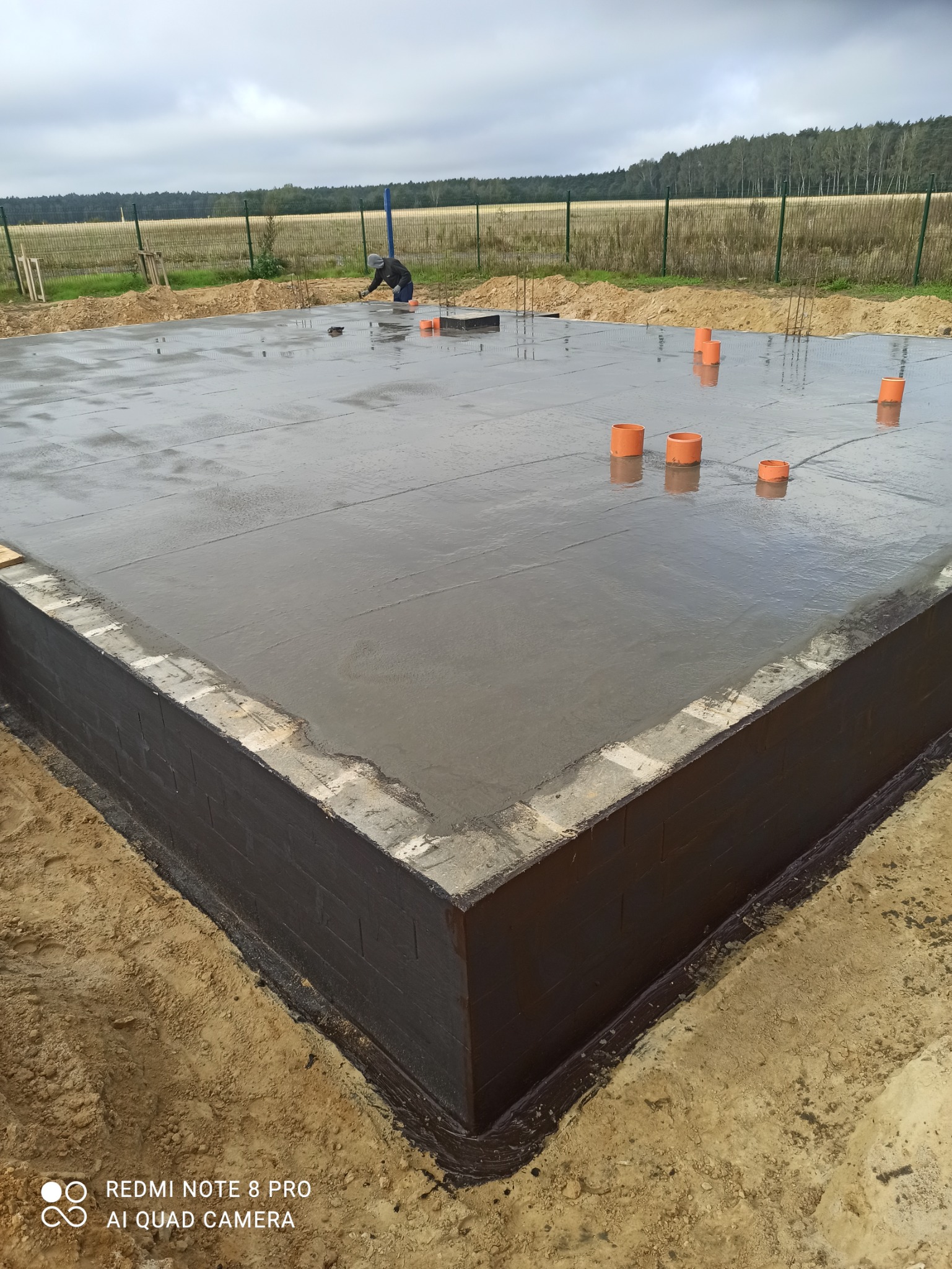 Świeżo wylana, mokra płyta fundamentowa z zabezpieczeniem przeciwwilgociowym na ścianach, widoczne pionowe pręty zbrojeniowe i pomarańczowe rury kanalizacyjne. W tle krajobraz polny pod pochmurnym...