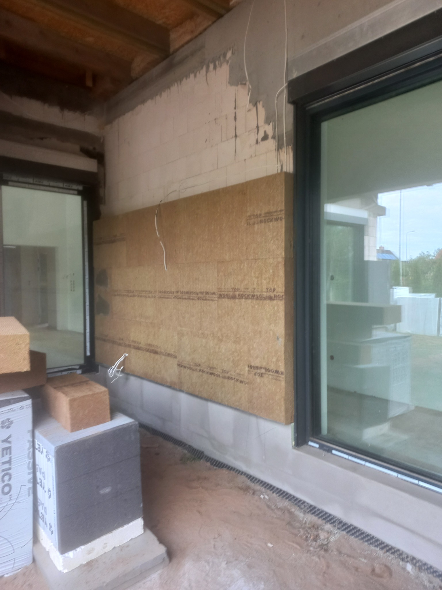 Fragment ściany zewnętrznej budynku w trakcie ocieplania wełną mineralną, widoczne okno z czarną ramą i ułożone obok bloki izolacyjne YETICO.