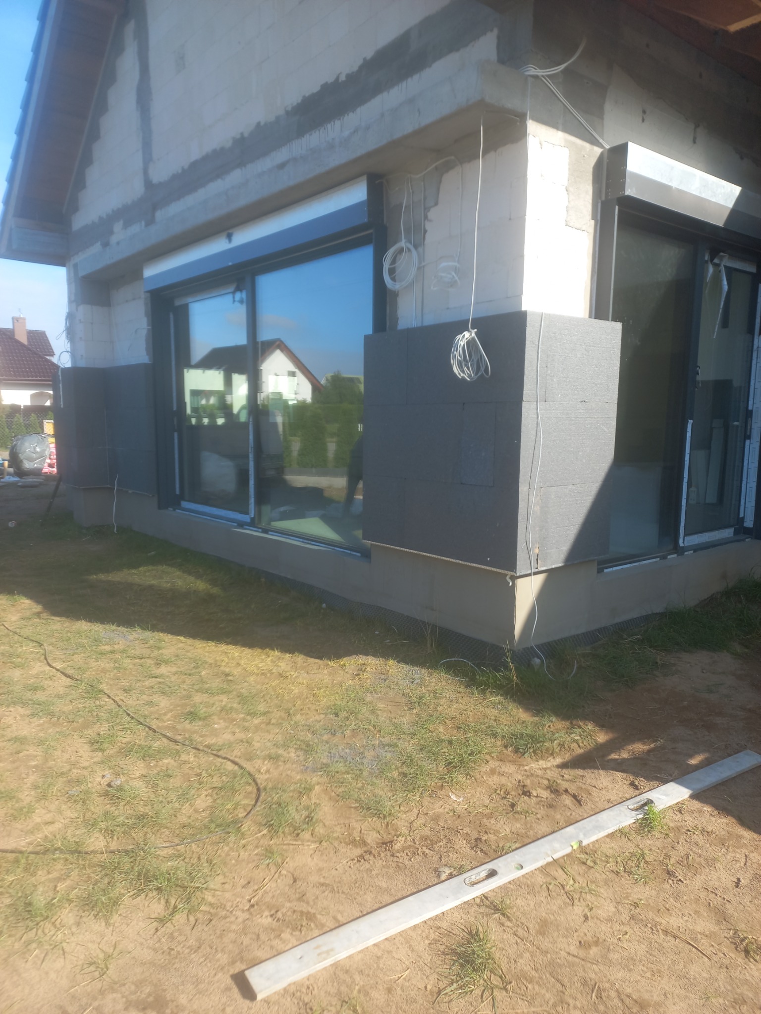 Fragment elewacji domu w trakcie ocieplania: przy oknach widoczne szare płyty izolacyjne, wystające kable, w tle okna z odbiciem otoczenia, na pierwszym planie trawa i poziomica.