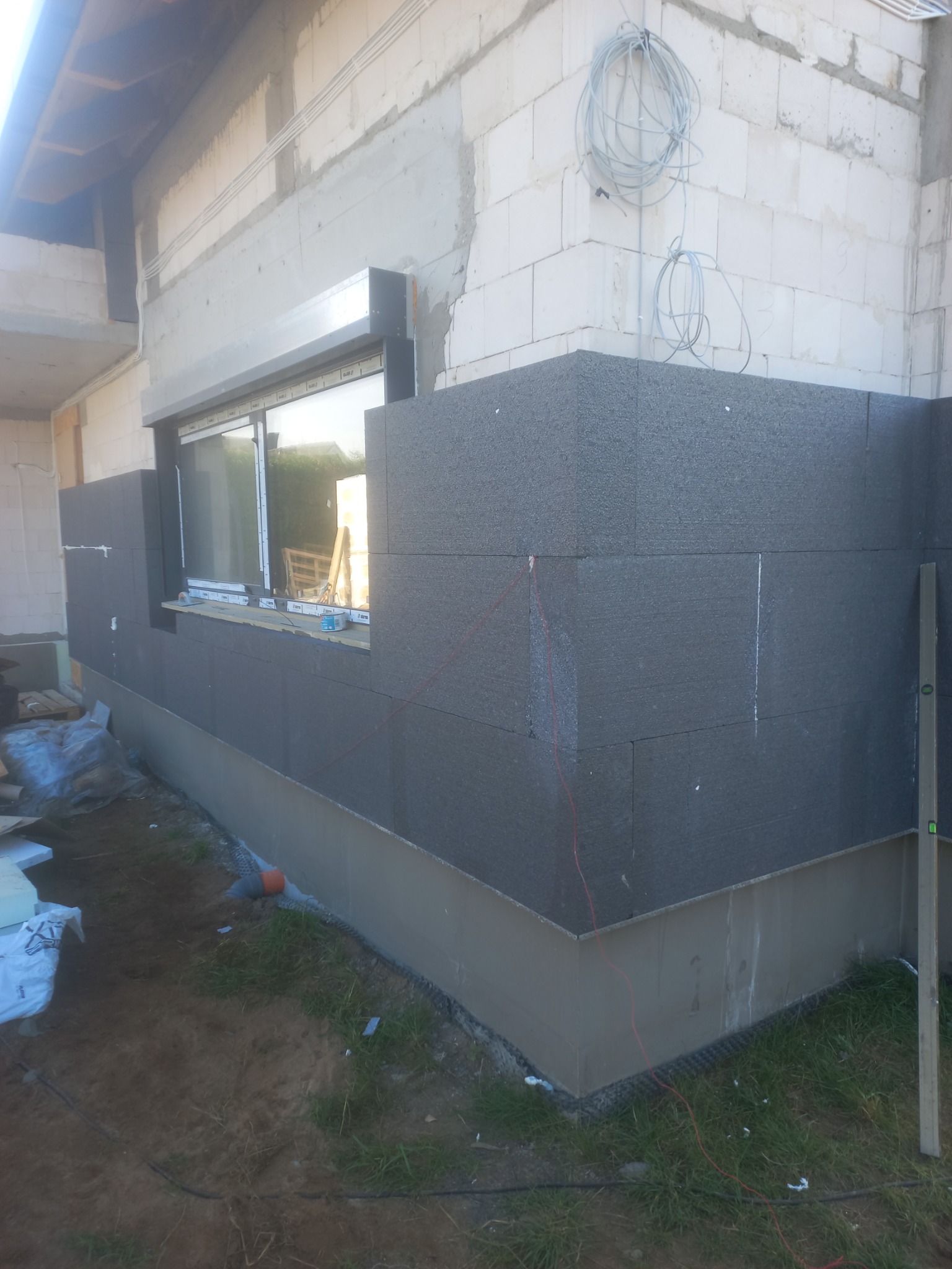 Ocieplanie ściany zewnętrznej budynku z szarego styropianu, widoczna poziomica, przewody elektryczne i okno z odbiciem krajobrazu.