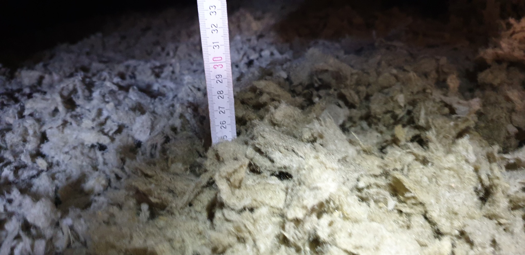 Pomiar grubości warstwy izolacyjnej wełny mineralnej na poddaszu za pomocą rozsuwanej miarki.
