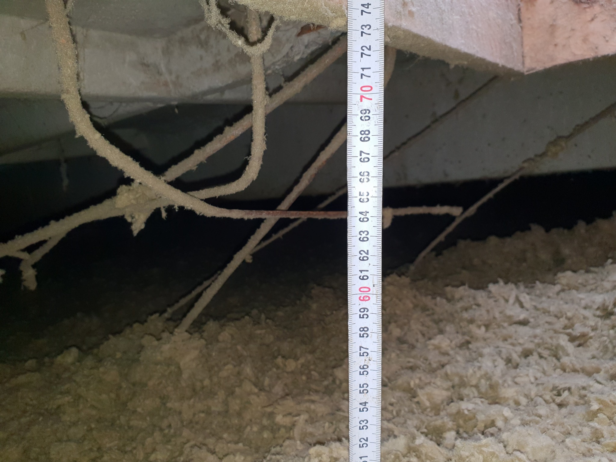 Pomiar grubości izolacji poddasza za pomocą rozwijanej miarki, widoczne przewody i elementy konstrukcyjne dachu.