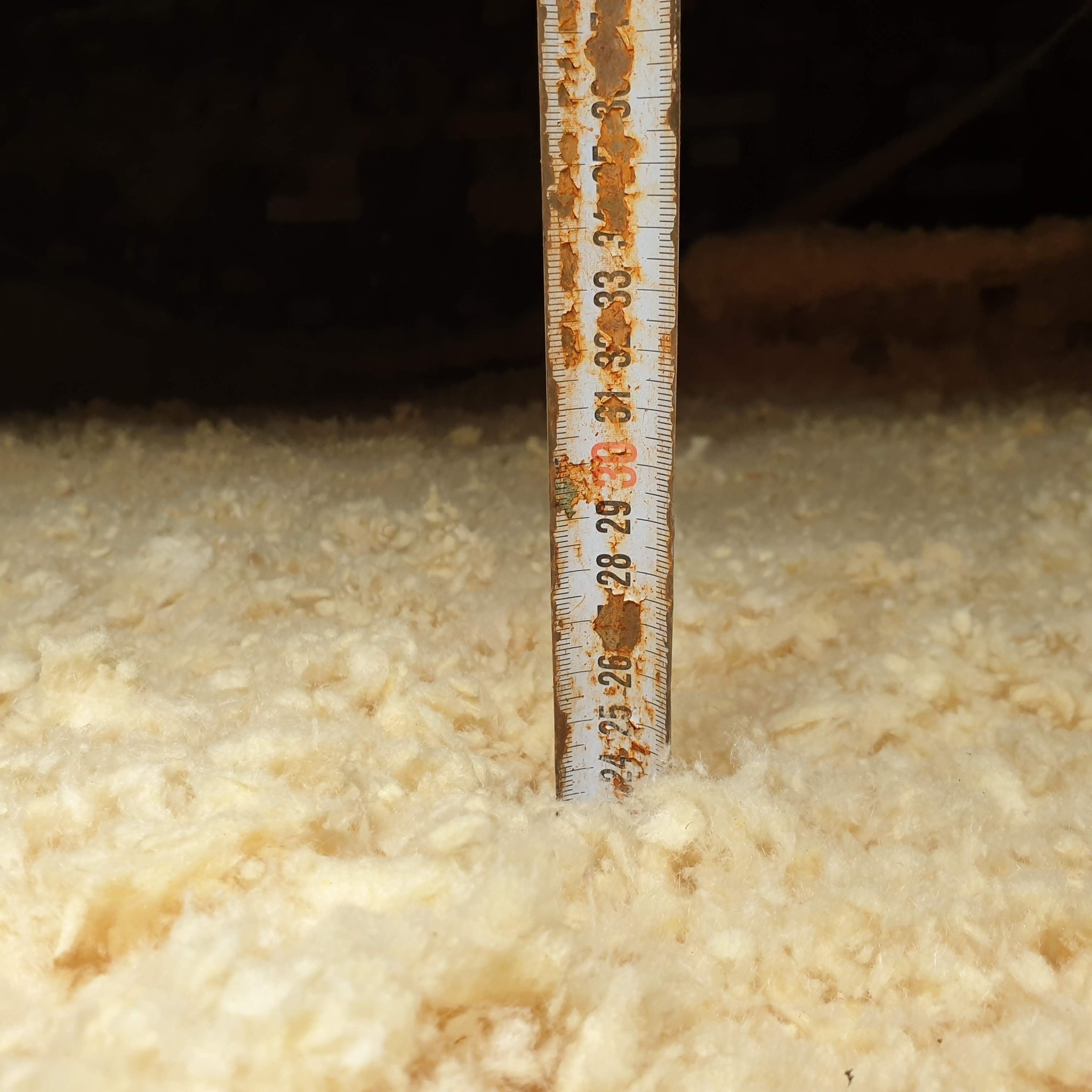 Pomiar grubości warstwy izolacyjnej z wełny mineralnej za pomocą zardzewiałej miarki w poddaszu.