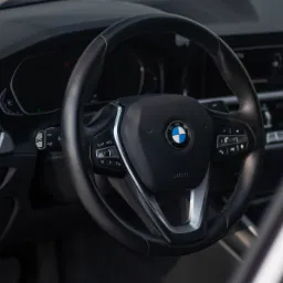 Wnętrze samochodu BMW z czarną skórzaną kierownicą, widoczne logo marki, deska rozdzielcza i elementy sterowania