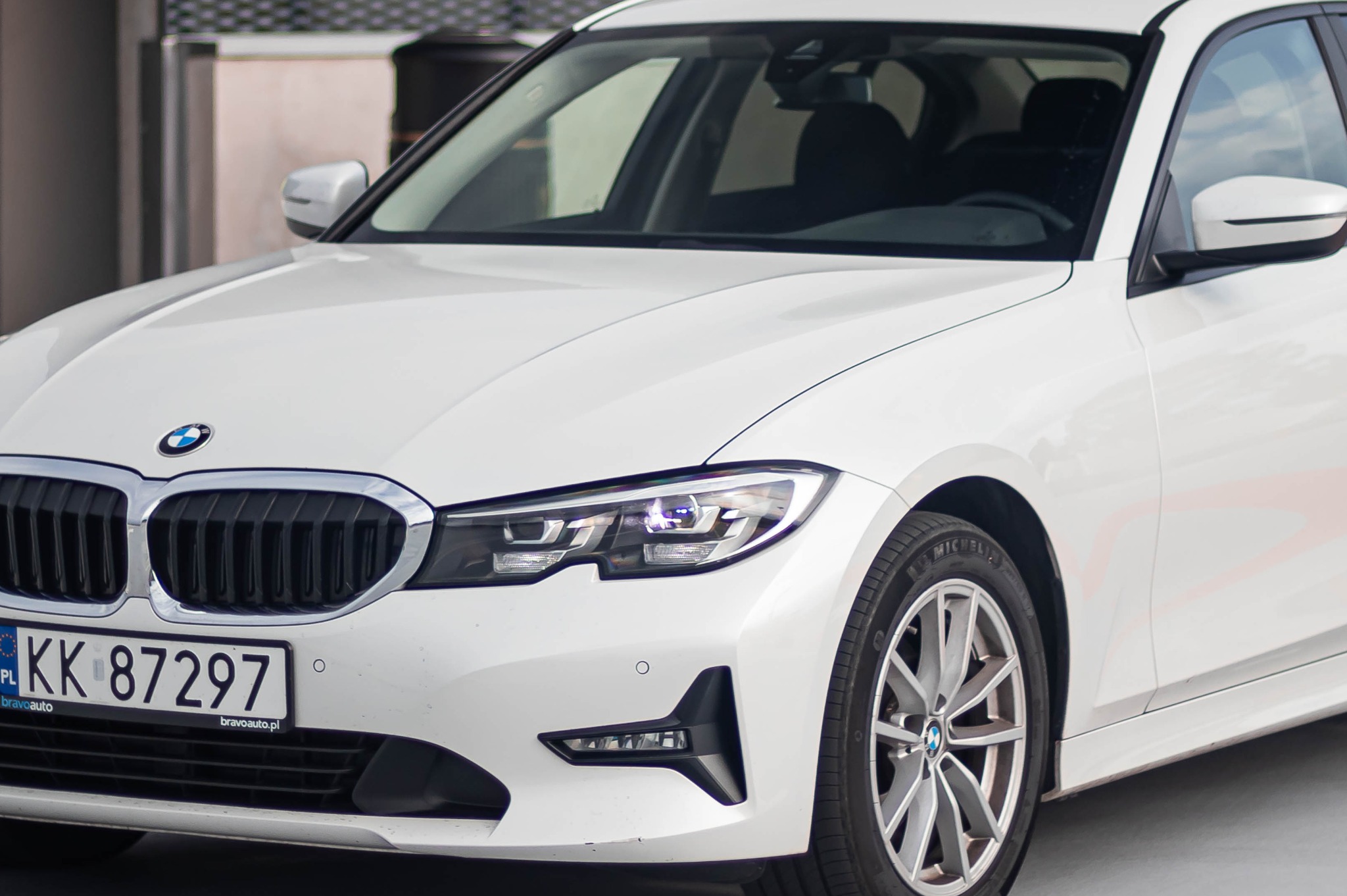 Białe BMW serii 3 z widoczną tablicą rejestracyjną 'KK 87297' i logo Bravoauto.pl na naklejce.