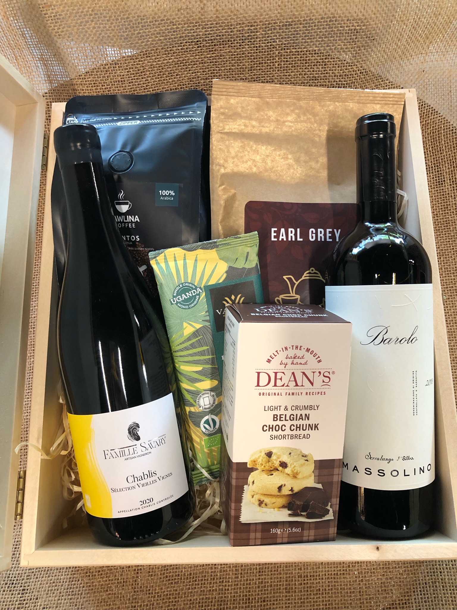 Otwarty drewniany kosz prezentowy wypełniony butelkami wina Chablis i Barolo, kawą ziarnistą, herbatą Earl Grey, ciasteczkami belgijskimi i tubą z przekąską, całość na jutowym tle.