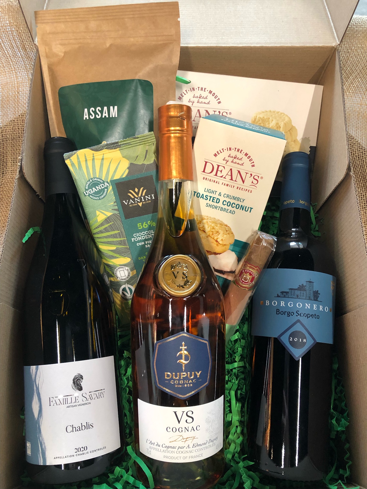 Elegancki kosz prezentowy z Warszawy, zawierający butelki wina Chablis i Borgonero, koniak Dupuy VS, kawę Assam, czekoladę Vanini z Ugandy, ciasteczka Dean's i cygaro, ułożone na zielonym...