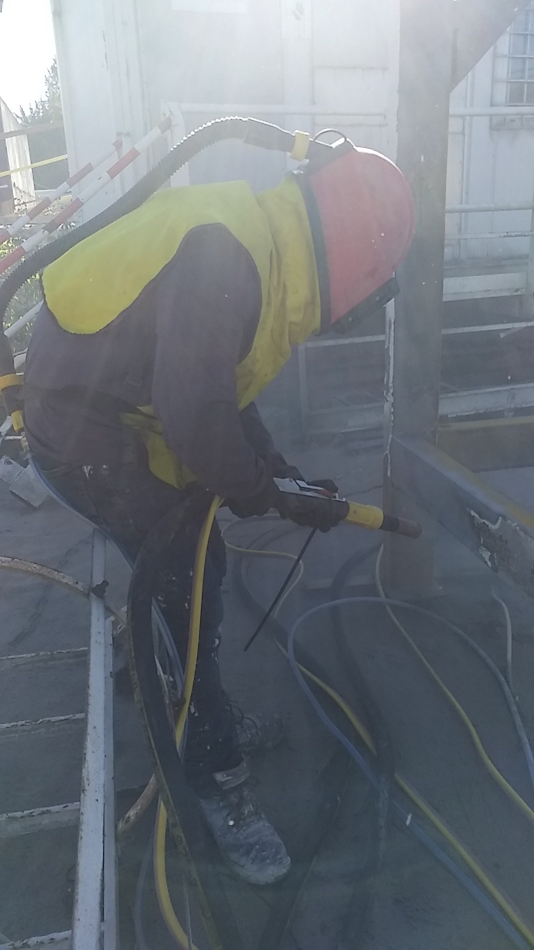 Pracownik w kombinezonie ochronnym, z maską i żółtą kamizelką, używa pistoletu do czyszczenia powierzchni metalowej, widoczne węże i pył.