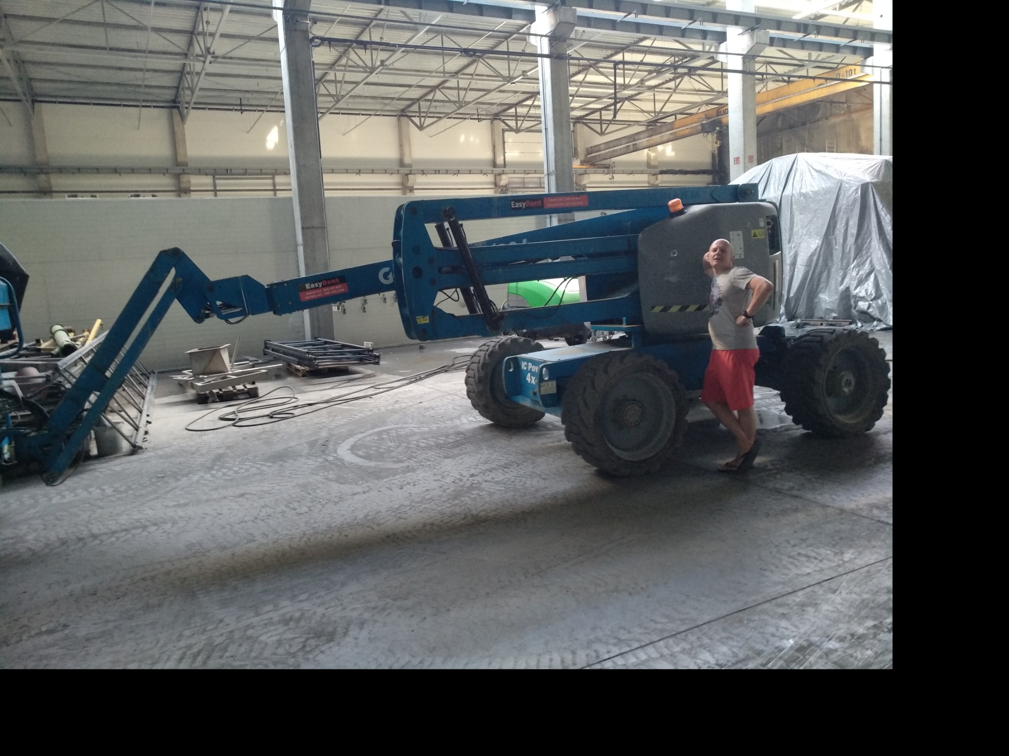 Mężczyzna w czerwonych spodenkach opiera się o niebieski podnośnik koszowy EasyRent w hali przemysłowej z betonową posadzką i metalową konstrukcją dachu.