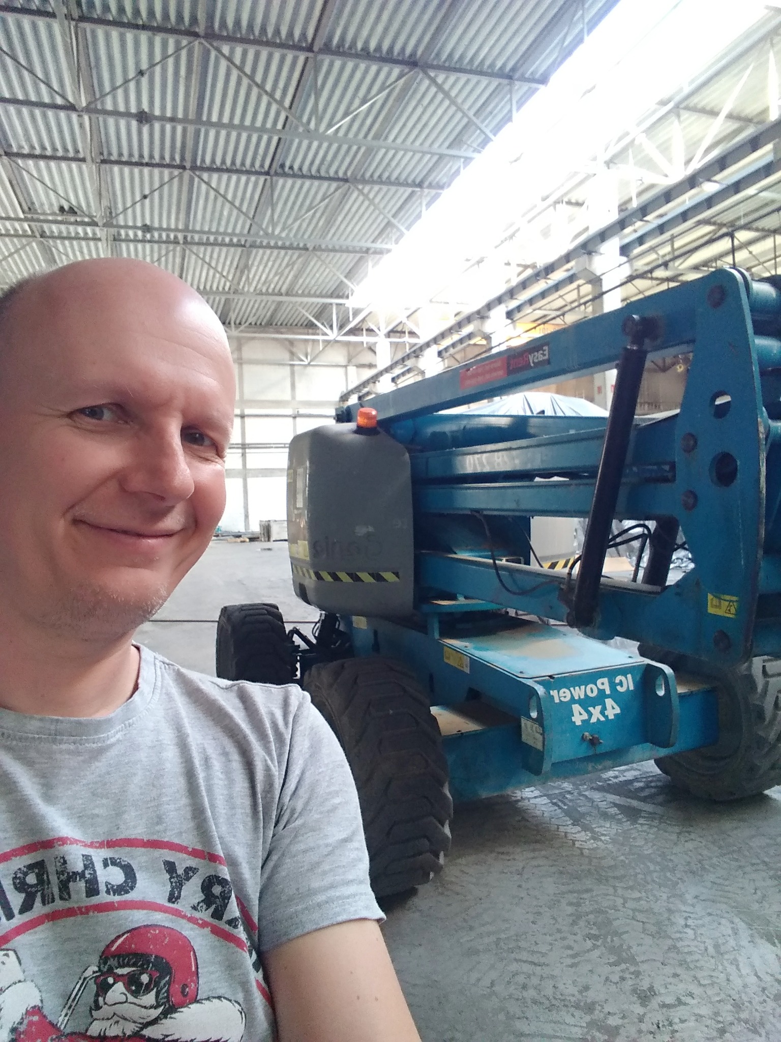 Uśmiechnięty mężczyzna robi selfie na tle podnośnika koszowego w hali przemysłowej z blachy falistej.