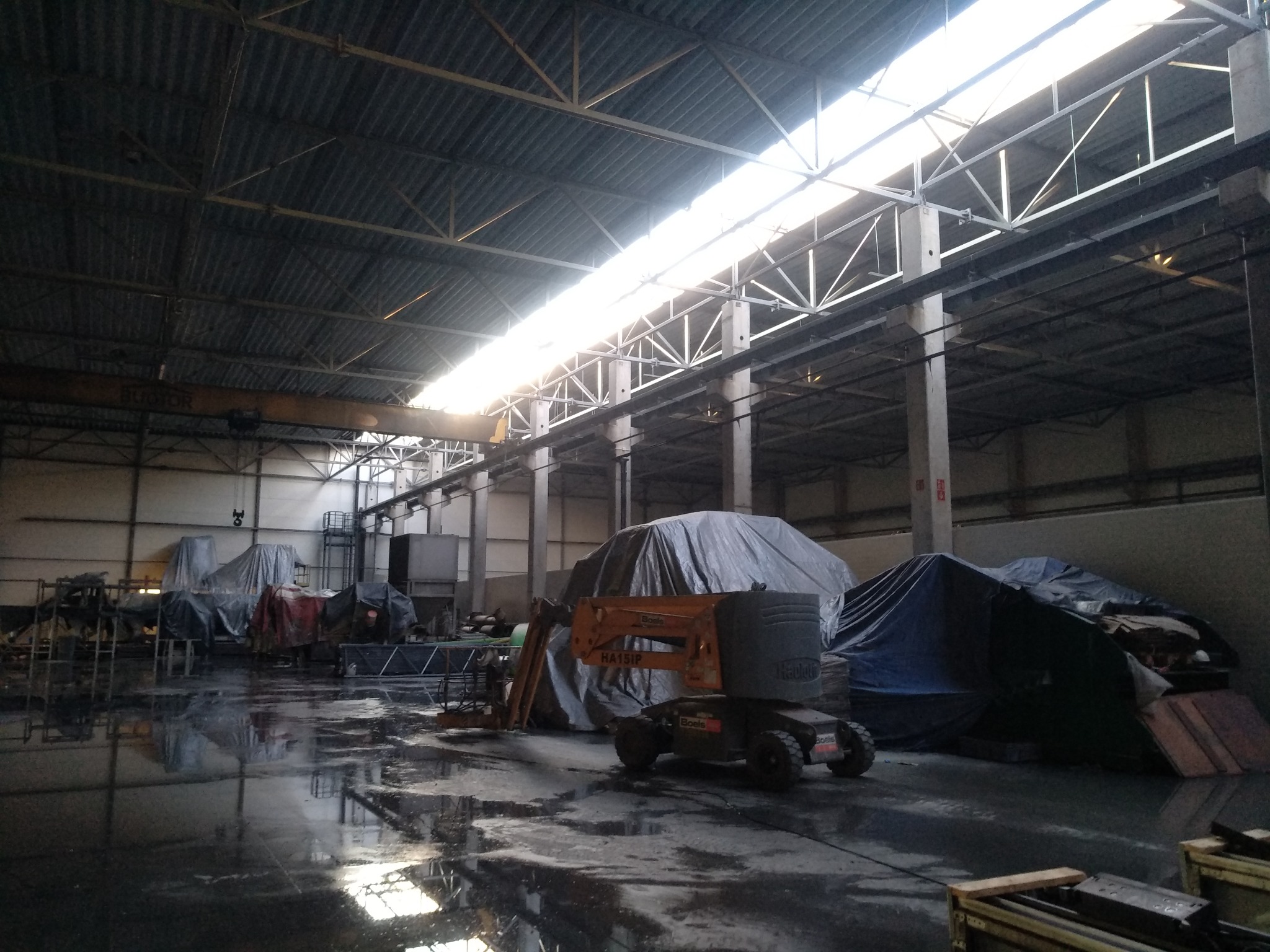 Wnętrze hali przemysłowej z podnośnikiem koszowym, materiałami budowlanymi pod plandekami i mokrą posadzką odbijającą światło z okien dachowych.