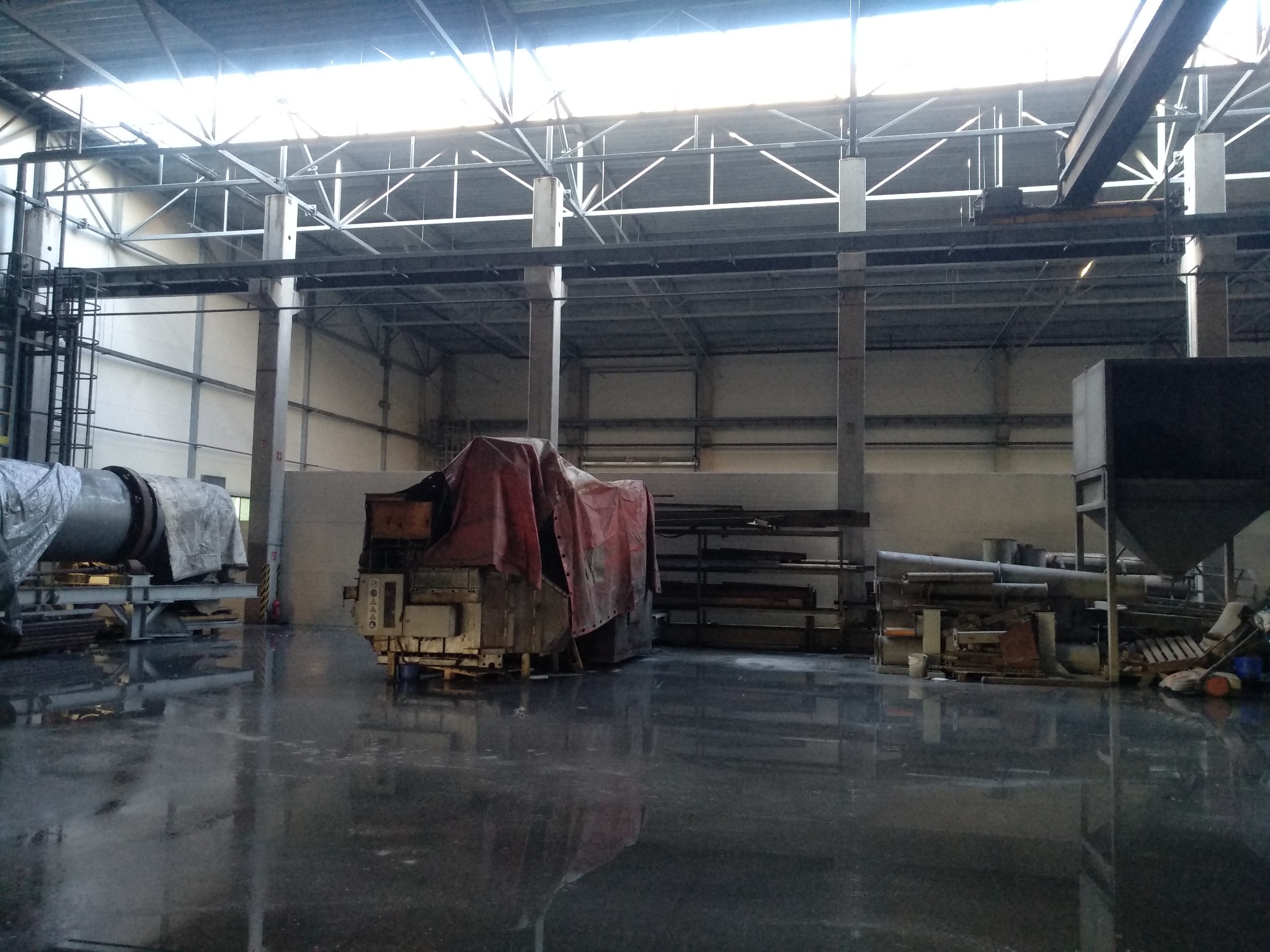 Wnętrze hali przemysłowej z maszynami przykrytymi plandekami, posadzka z refleksami świetlnymi, widoczna konstrukcja dachu i suwnica.