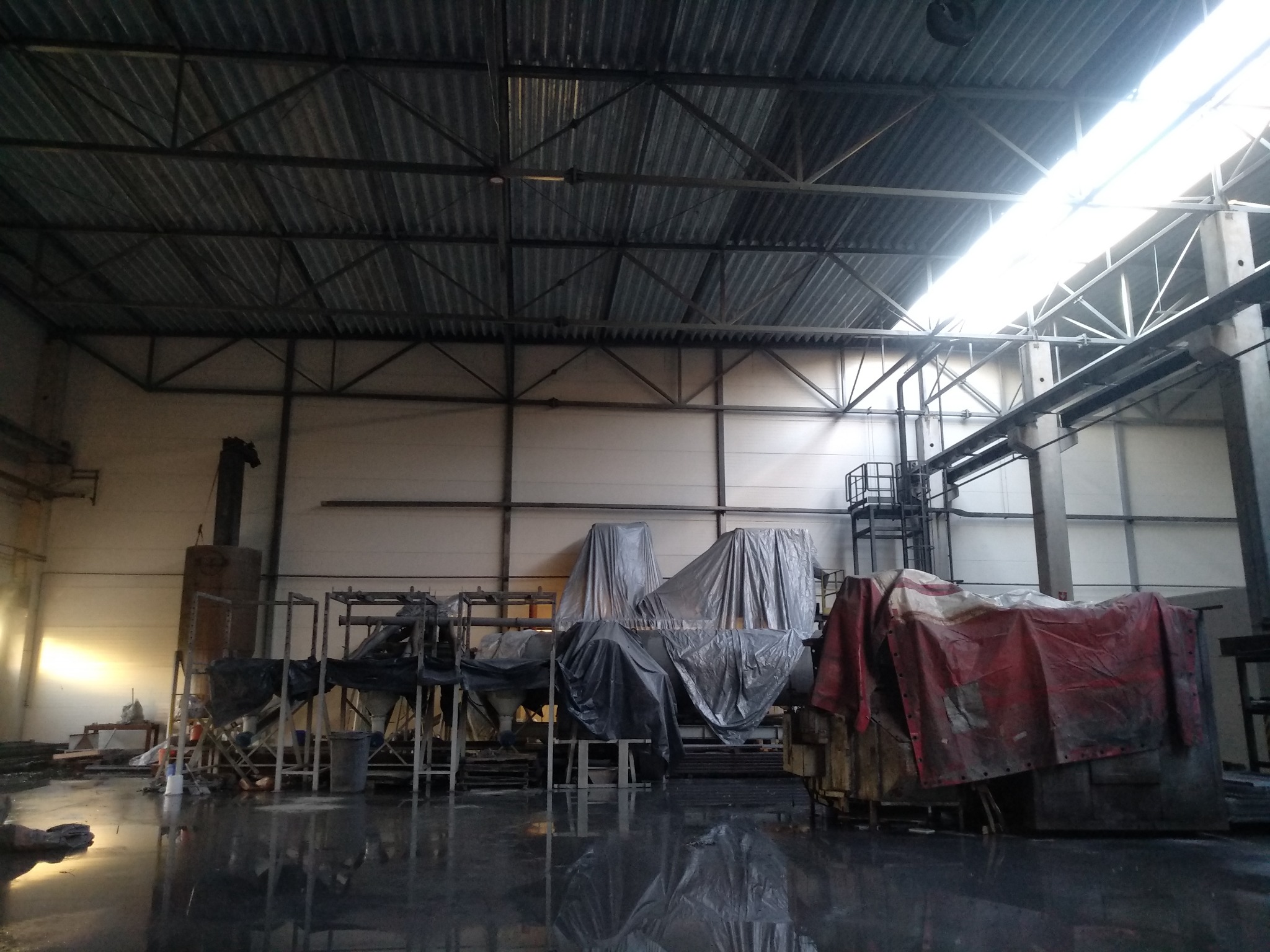 Wnętrze hali przemysłowej z metalową konstrukcją dachu i maszynami częściowo zakrytymi plandekami, mokra posadzka odbija światło wpadające przez okna dachowe.