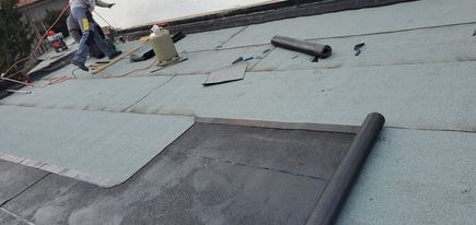 Prace na dachu: układanie papy termozgrzewalnej na warstwie izolacyjnej, widoczny palnik, rolki papy i pracownicy w tle.