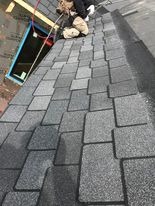 Pracownik w trakcie montażu bitumicznych płytek dachowych na skośnym dachu, widoczny fragment okna dachowego i narzędzia.