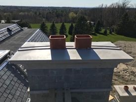 Wykończony komin z dwoma ceramicznymi wkładami, widok z góry na tle zielonego krajobrazu, fragment dachu pokrytego szarą dachówką widoczny w lewym górnym rogu.