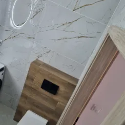 Nowoczesna łazienka z białymi płytkami imitującymi marmur z złotymi żyłkami, drewnianą zabudową stelaża WC z czarnym przyciskiem, okrągłym lustrem z podświetleniem LED i białym sedesem wiszącym.