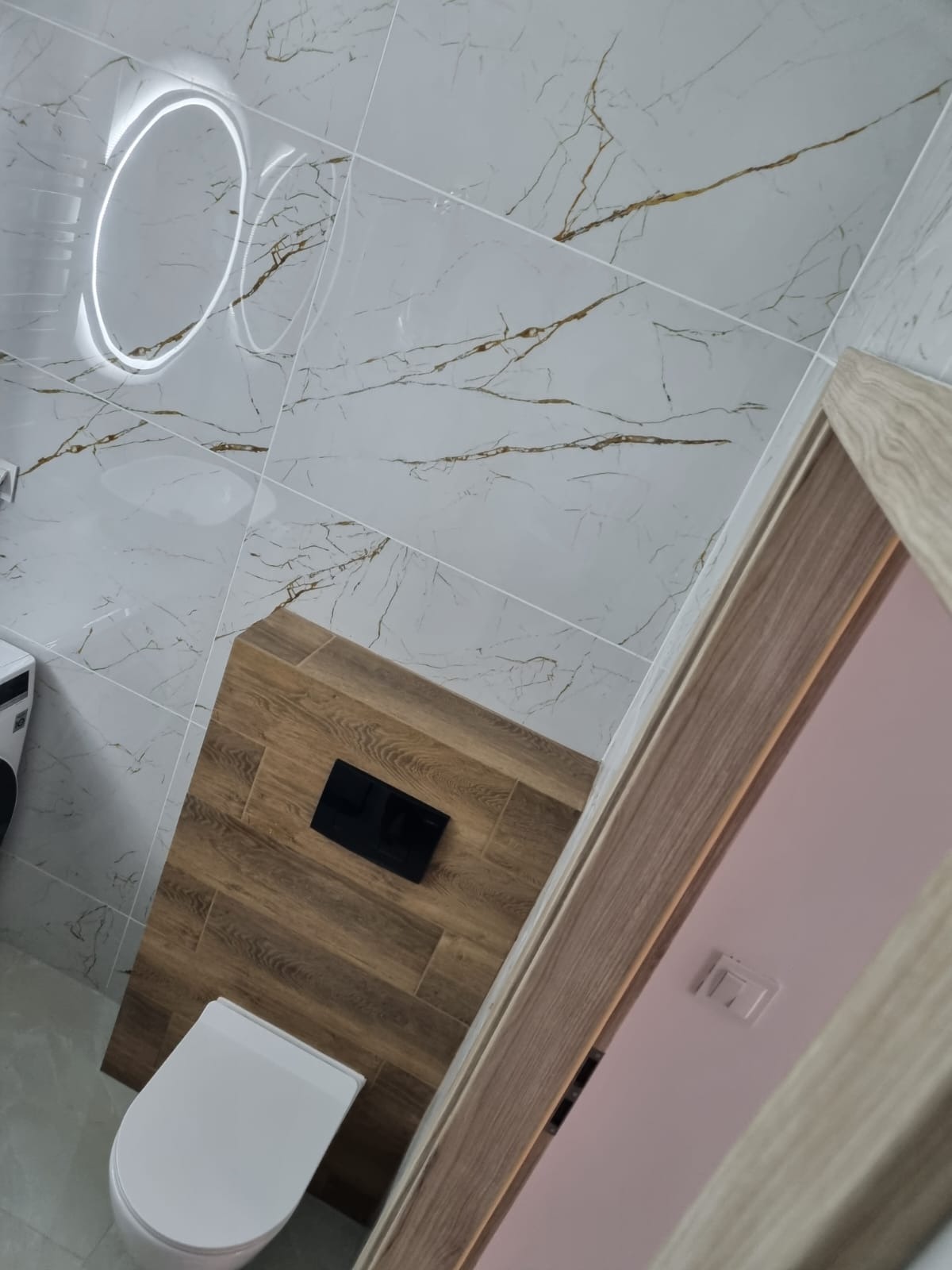 Nowoczesna łazienka z białymi płytkami imitującymi marmur z złotymi żyłkami, drewnianą zabudową stelaża WC z czarnym przyciskiem, okrągłym lustrem z podświetleniem LED i białym sedesem wiszącym.