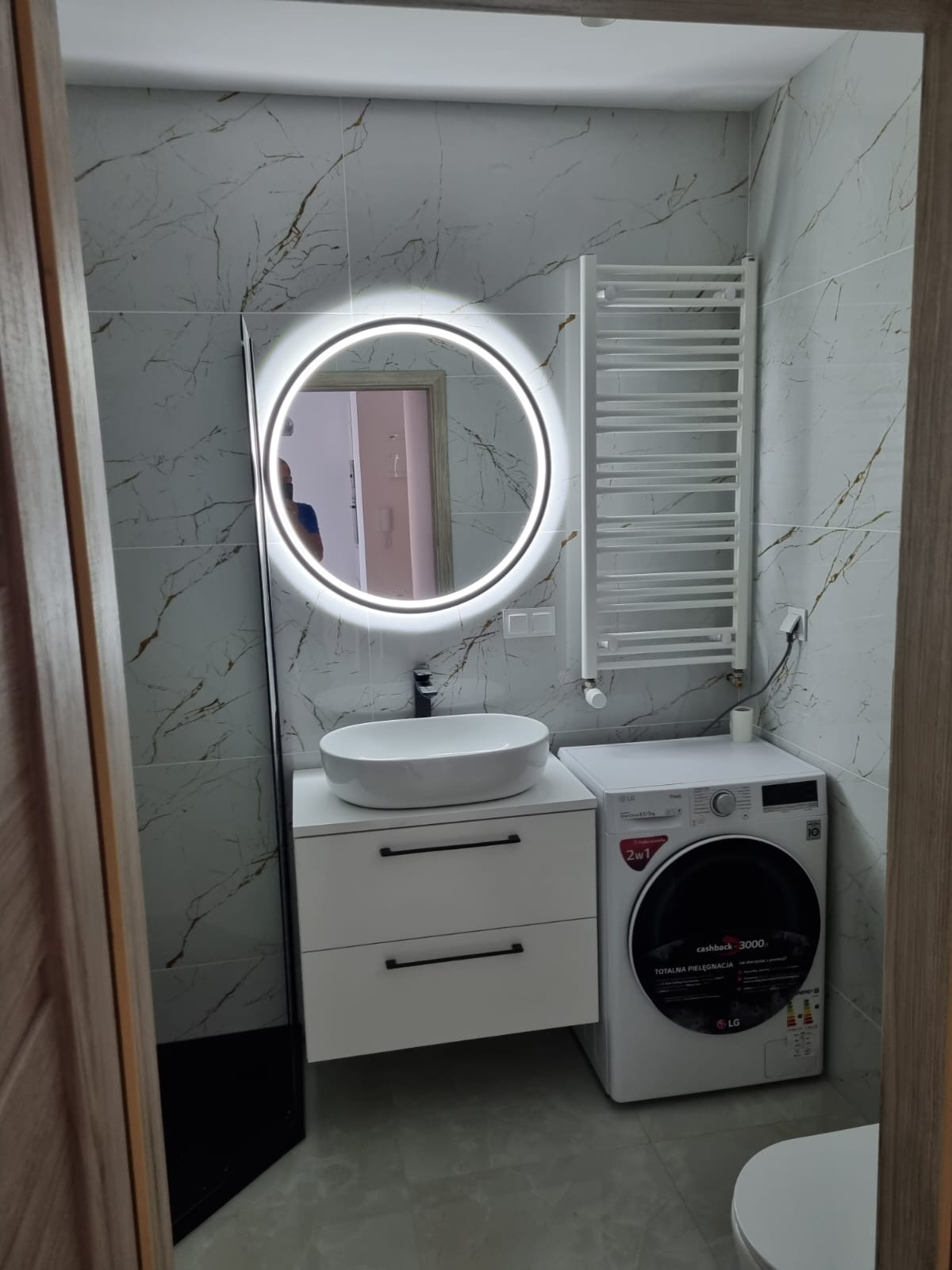 Nowoczesna łazienka z marmurowymi płytkami, okrągłym lustrem z podświetleniem LED, białą umywalką nablatową, szafką z szufladami, grzejnikiem drabinkowym i pralką LG