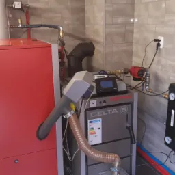 Instalacja kotła na paliwo stałe w kotłowni z szarą płytką, widoczny czerwony korpus kotła, szary kocioł z podajnikiem i elementami sterującymi, oraz orurowanie z zaworami i pompami.