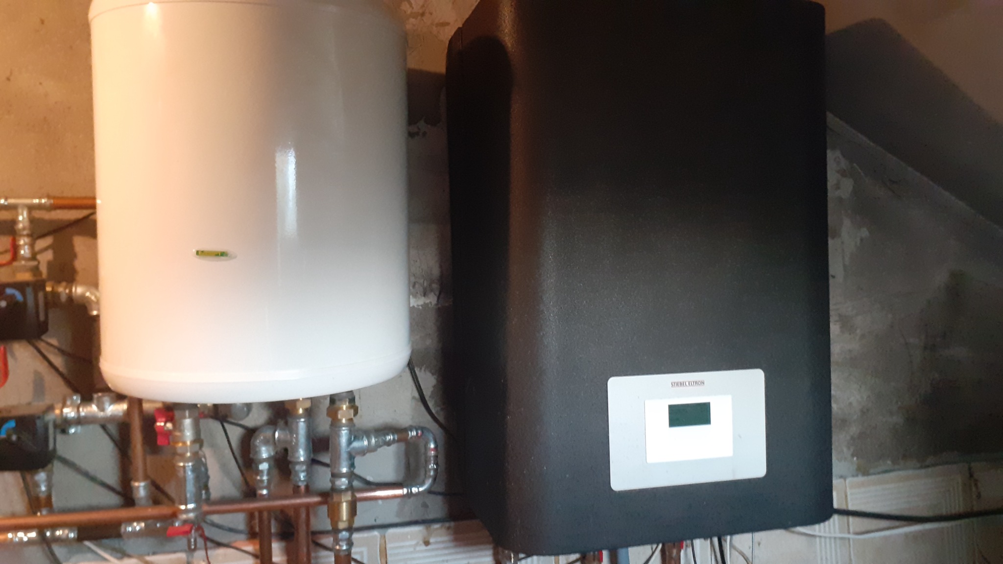 Biały zasobnik ciepłej wody i czarna jednostka centralna w kotłowni w Strzelnie, z widocznymi rurami miedzianymi i elementami instalacji.