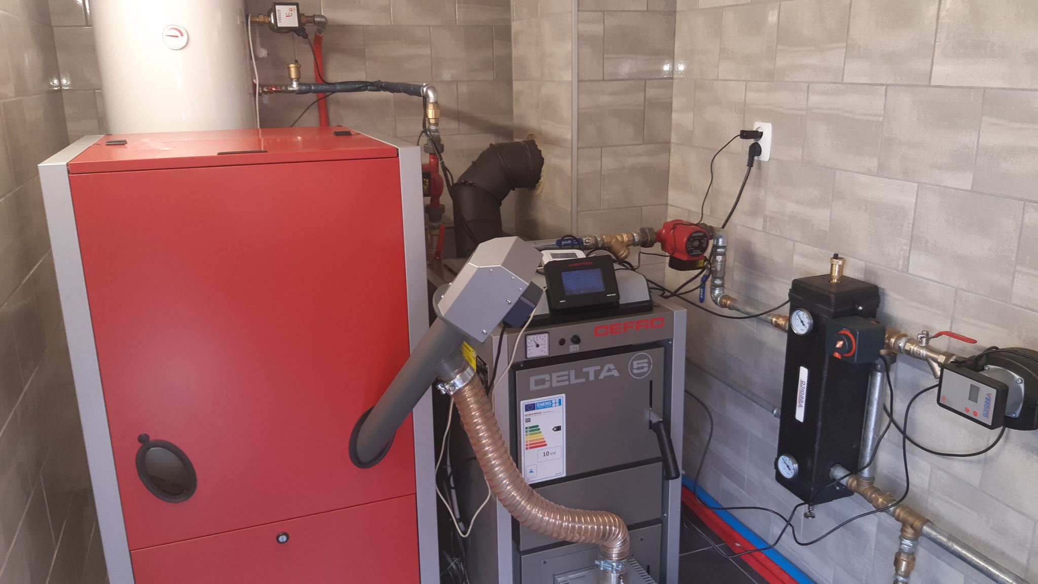 Instalacja kotła na paliwo stałe w kotłowni z szarą płytką, widoczny czerwony korpus kotła, szary kocioł z podajnikiem i elementami sterującymi, oraz orurowanie z zaworami i pompami.